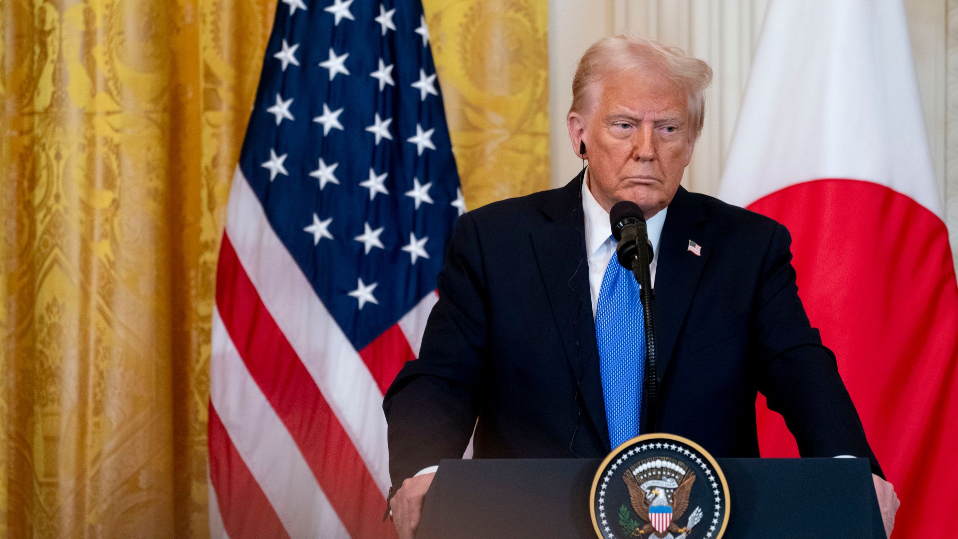 Man in dark suit and blue tie speaking at a podium with the presidential seal, with U.S. and Japan flags in the background against gold curtains.