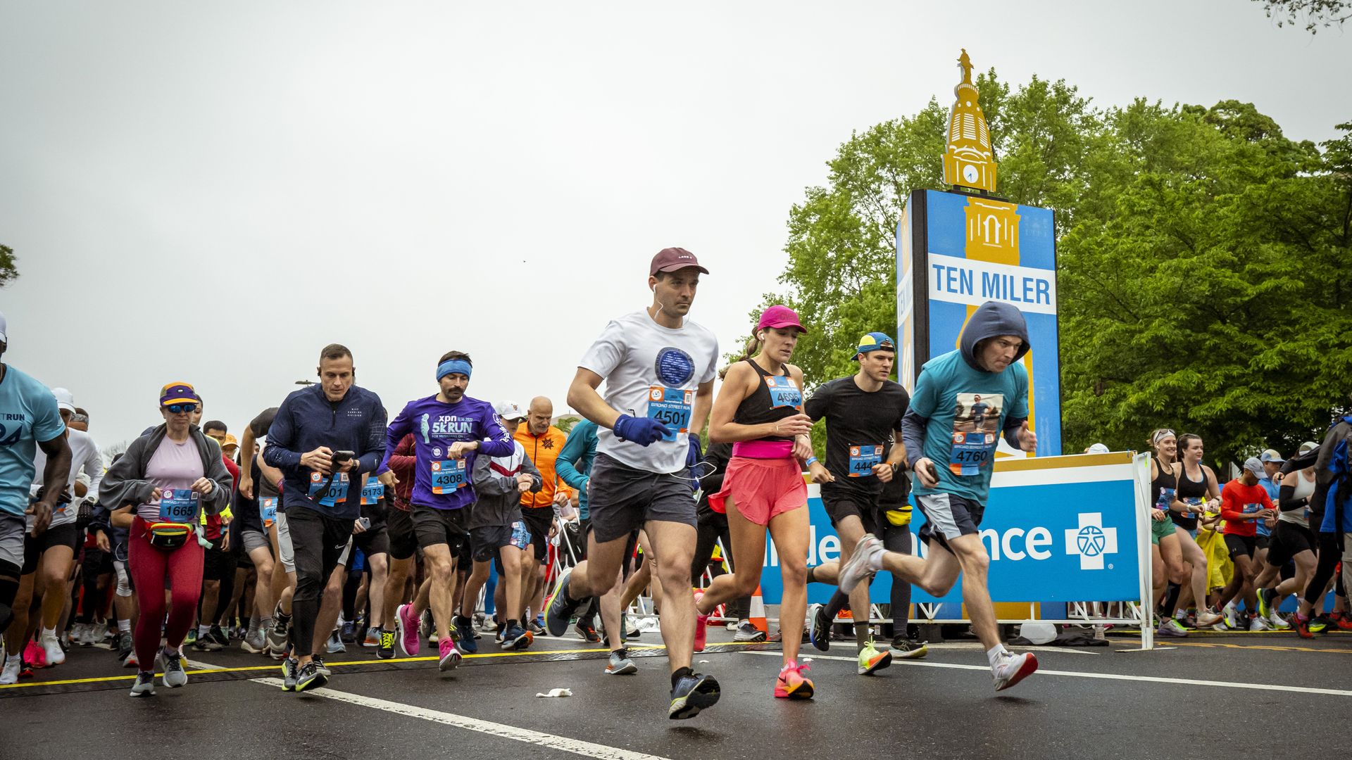 Runners at the start of a race, some in colorful athletic gear, passing a blue and yellow sign that reads "TEN MILER" with green trees in the background on an overcast day.