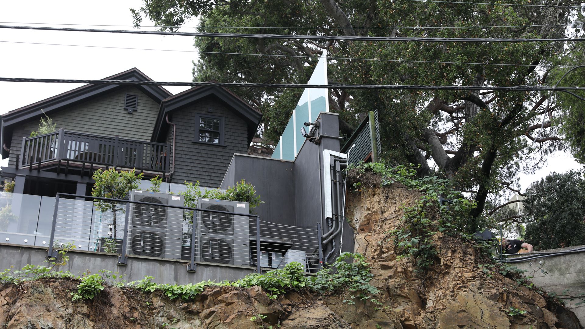 A gray two-story house with a wooden balcony sits on a rocky hillside. In front are three air-conditioning units behind a glass and metal fence; a large tree looms overhead with power lines across.