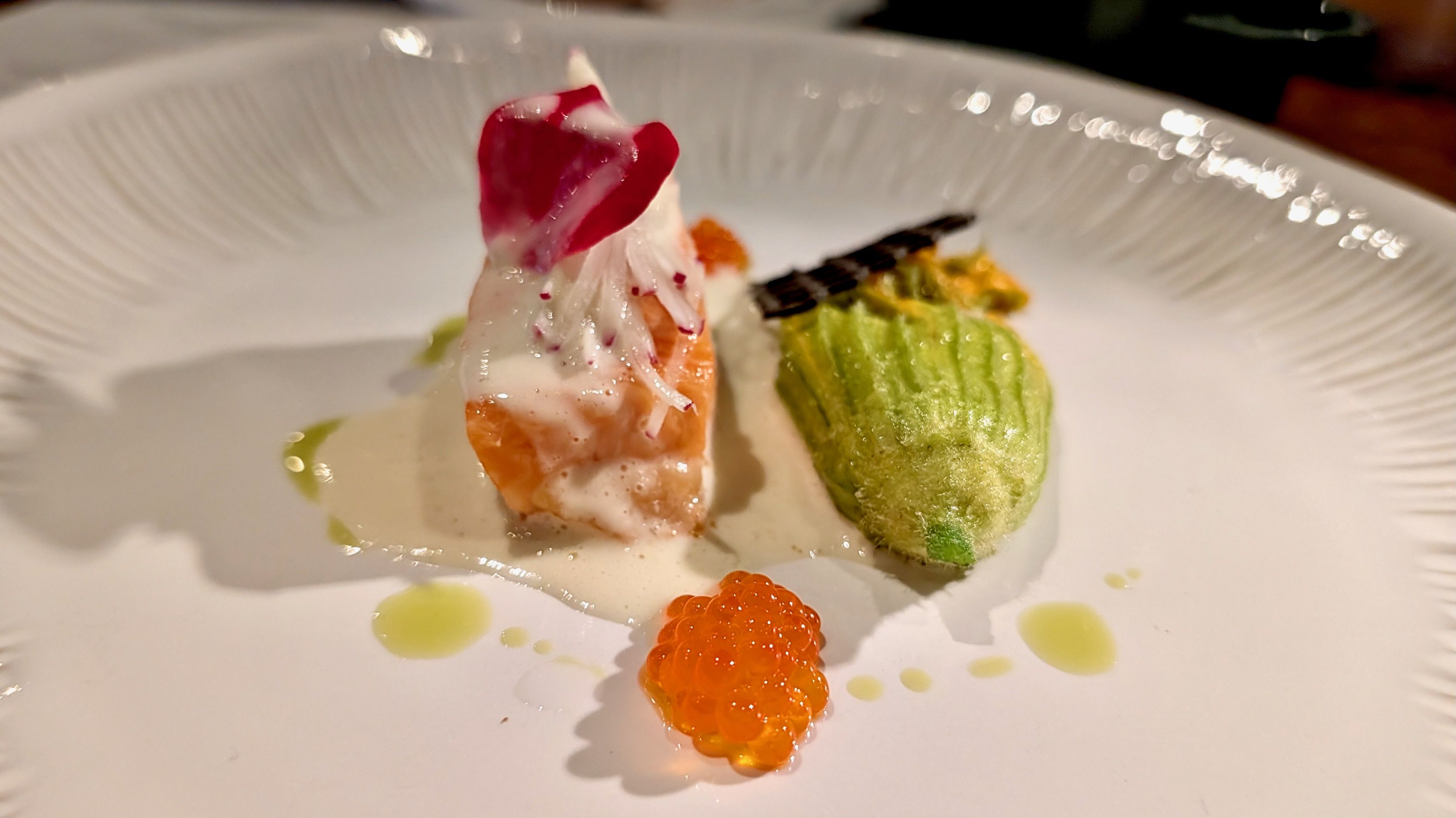 An elegant plated seafood appetizer: a shrimp with creamy white sauce and a red leaf garnish, a green vegetable with a brown-topped pattern, and small orange fish roe on a white plate.