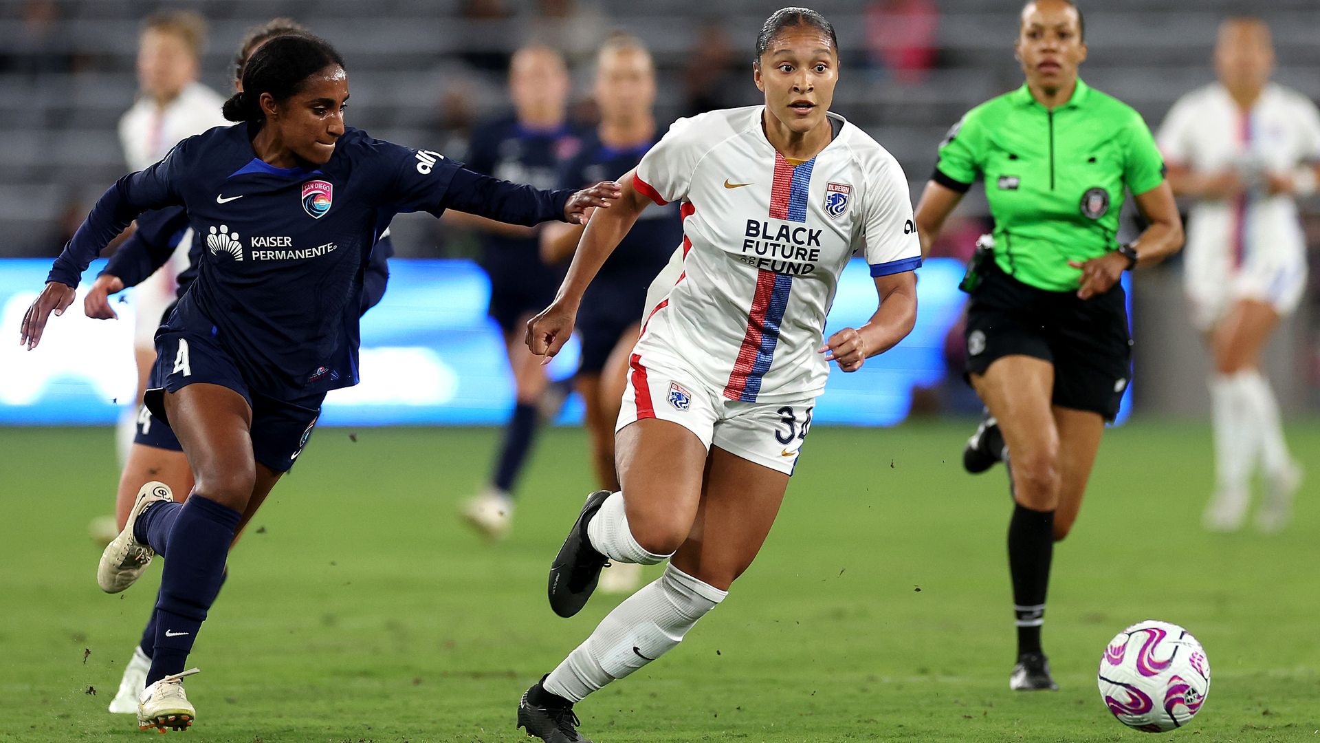 Two professional women's soccer players run after a ball on the field. 