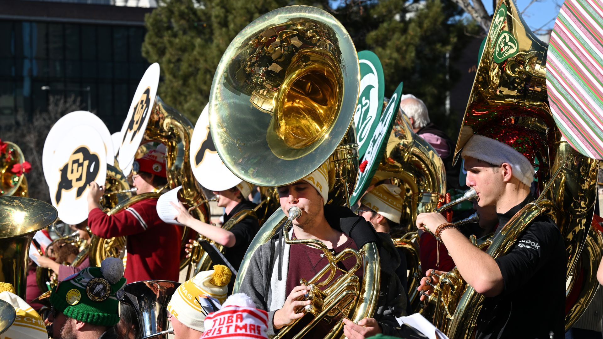 Several tuba players blow on large horns while wearing holiday attire. 