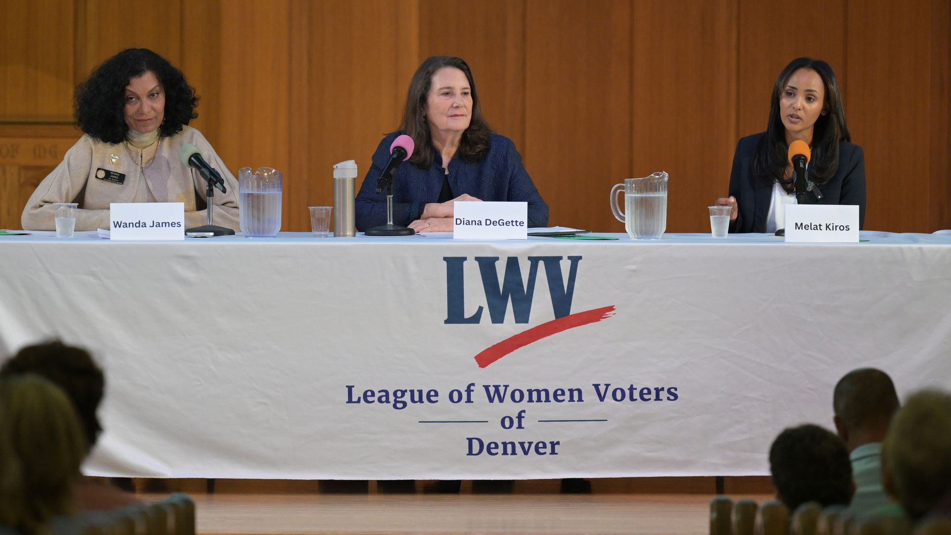 Panel discussion at the League of Women Voters of Denver: three women at a table with microphones; placards read Wanda James, Diana DeGette, Melat Kiros, against a wood-paneled backdrop.