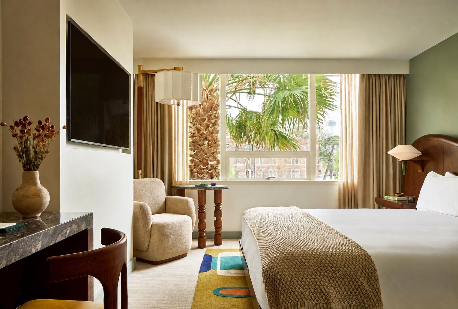 Hotel room with a large window view of palm trees. Bed with white linens and a beige knit throw; chair and small table by the window. TV on the wall, vase of dried flowers, desk nearby.