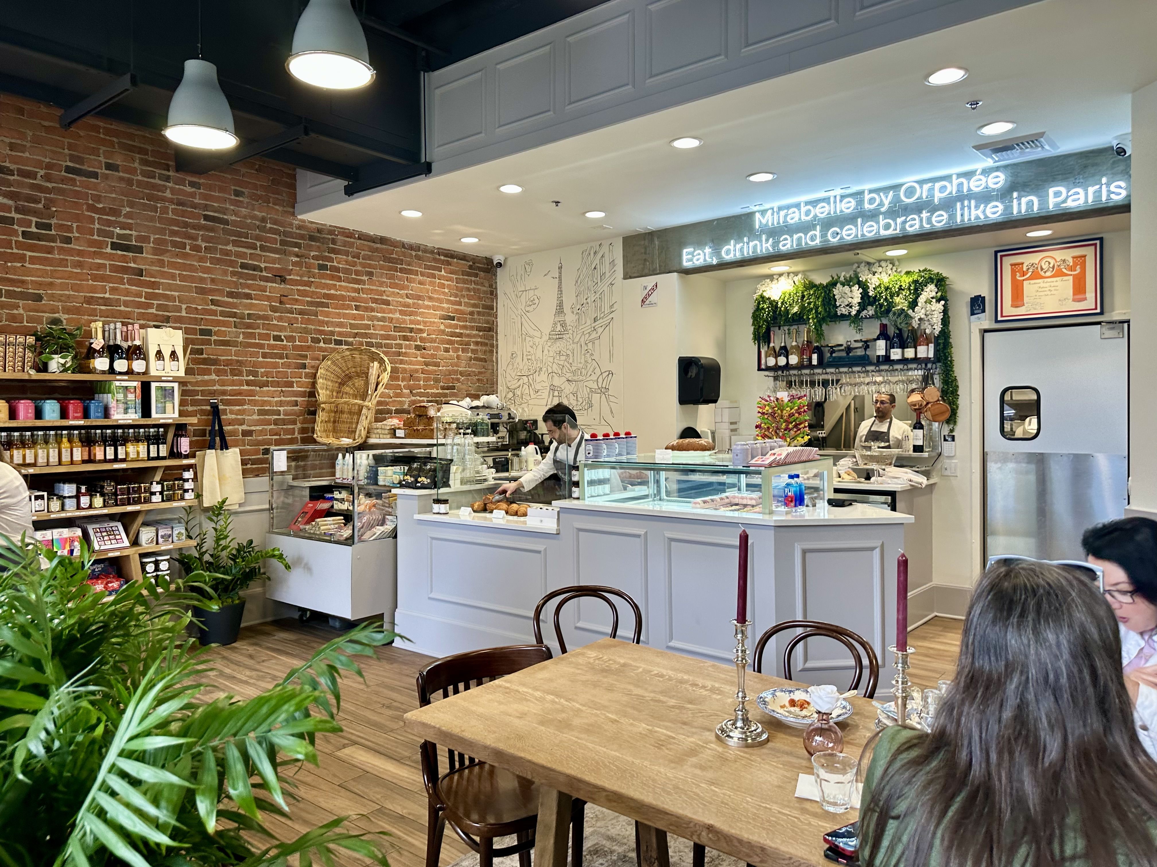 A look at the inside of Mirabelle by Orphée, with imported products on a shelf along a brick wall, a white wooden counter that holds the pastry case, and a window into the kitchen. People sit and eat at a wooden table while the owner Orphée Fouano works behind the counter. 