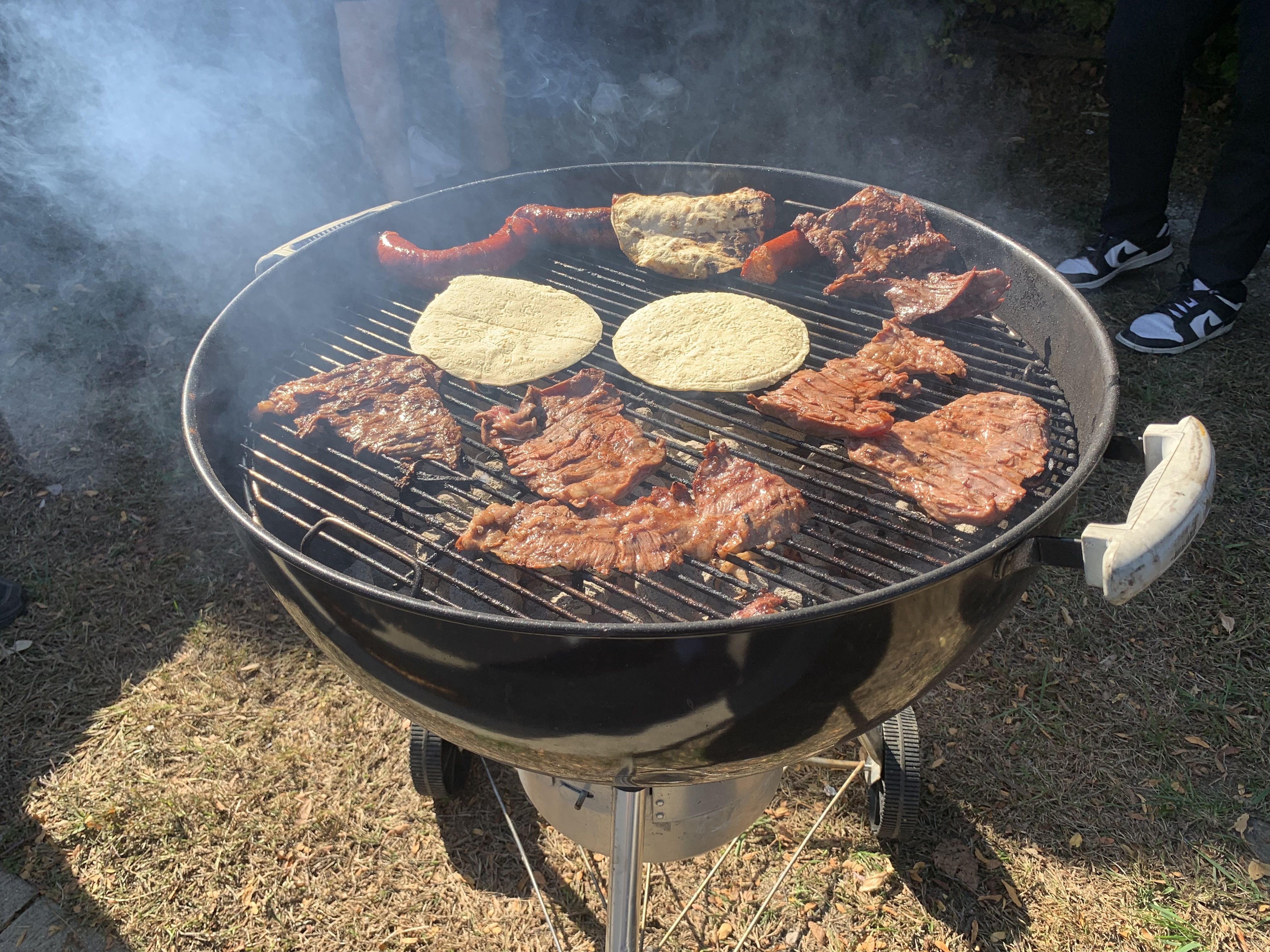 Round charcoal grill outdoors with multiple pieces of grilled meat and two tortillas on the grate; smoke rises as a person wearing sneakers stands nearby.