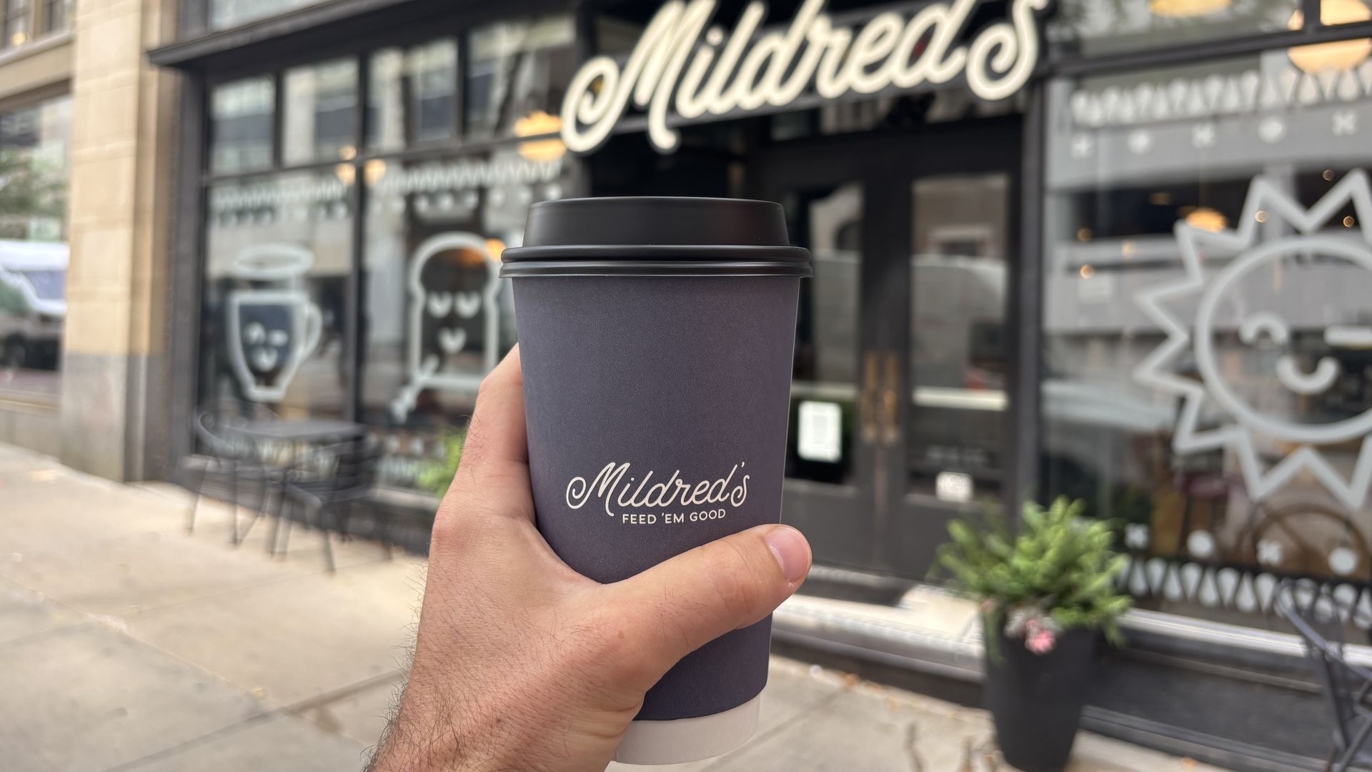 Hand holds a dark purple to-go coffee cup with a black lid in front of a storefront cafe. A large cursive sign reads Mildred's, with glass windows and outdoor seating visible.