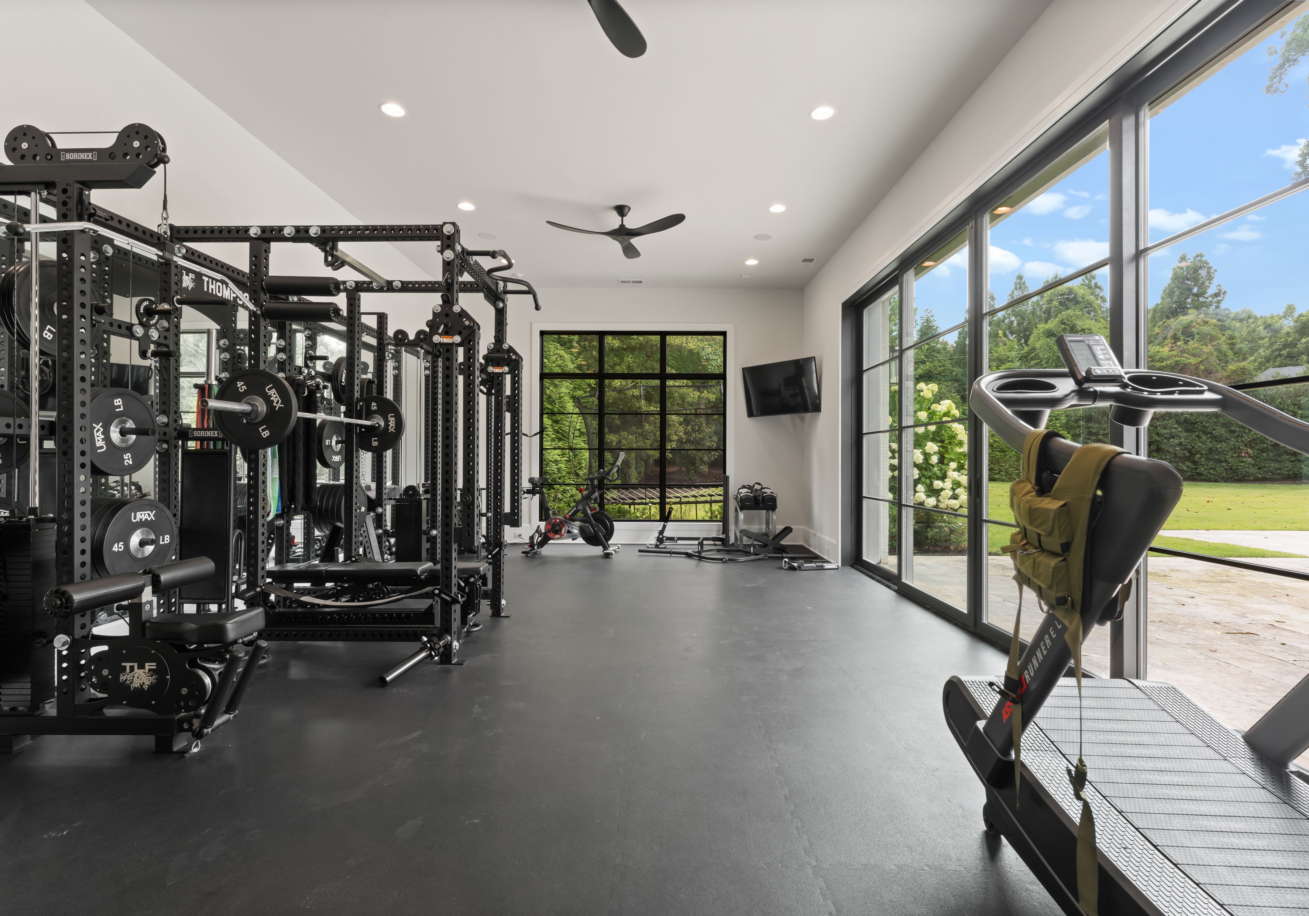 Bright home gym with black weight rack, treadmill, stationary bike, dumbbells, and large windows showing green yard and blue sky.