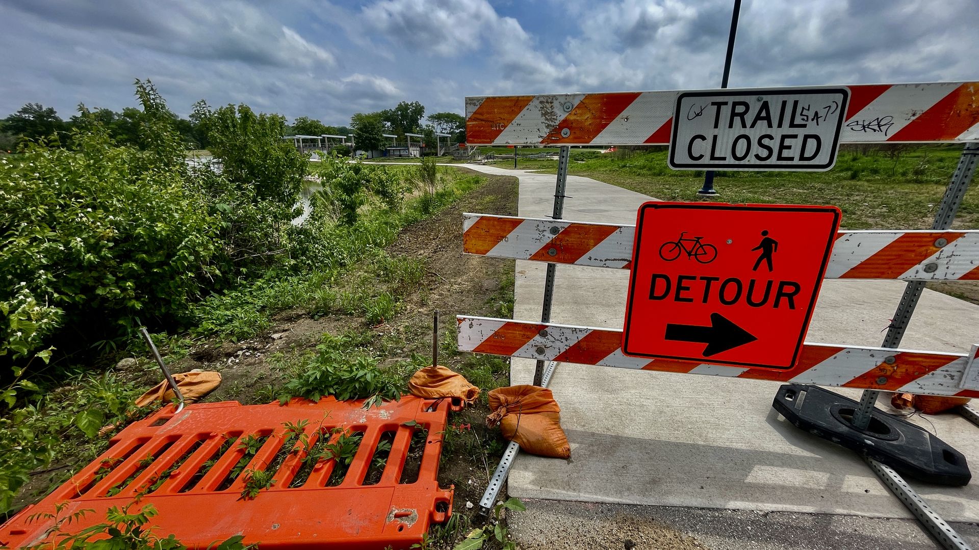 Orange-and-white barricades block a paved river trail, with a "TRAIL CLOSED" sign and a bright orange "DETOUR" sign showing bike and pedestrian icons; cloudy sky overhead.