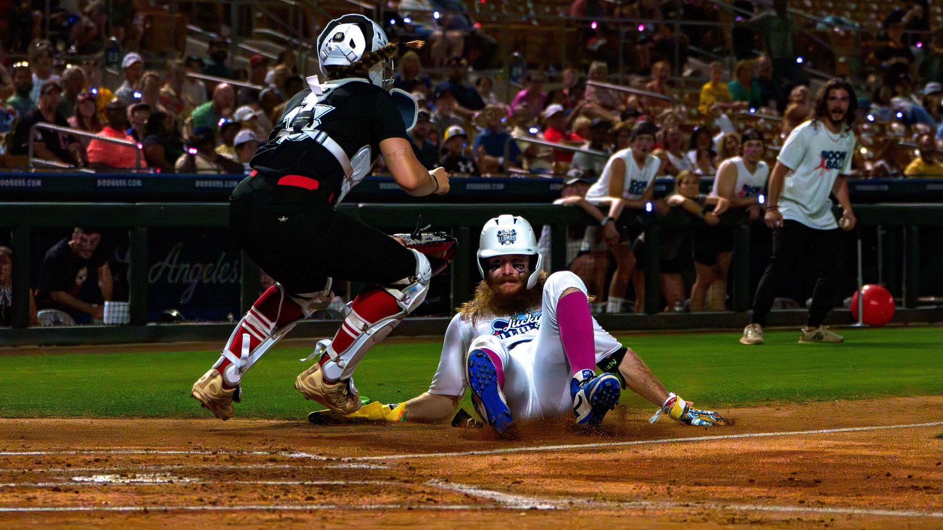 Bearded baserunner in white with a purple sleeve slides into home as a black-clad catcher leaps to tag; a crowded stadium watches from the stands.