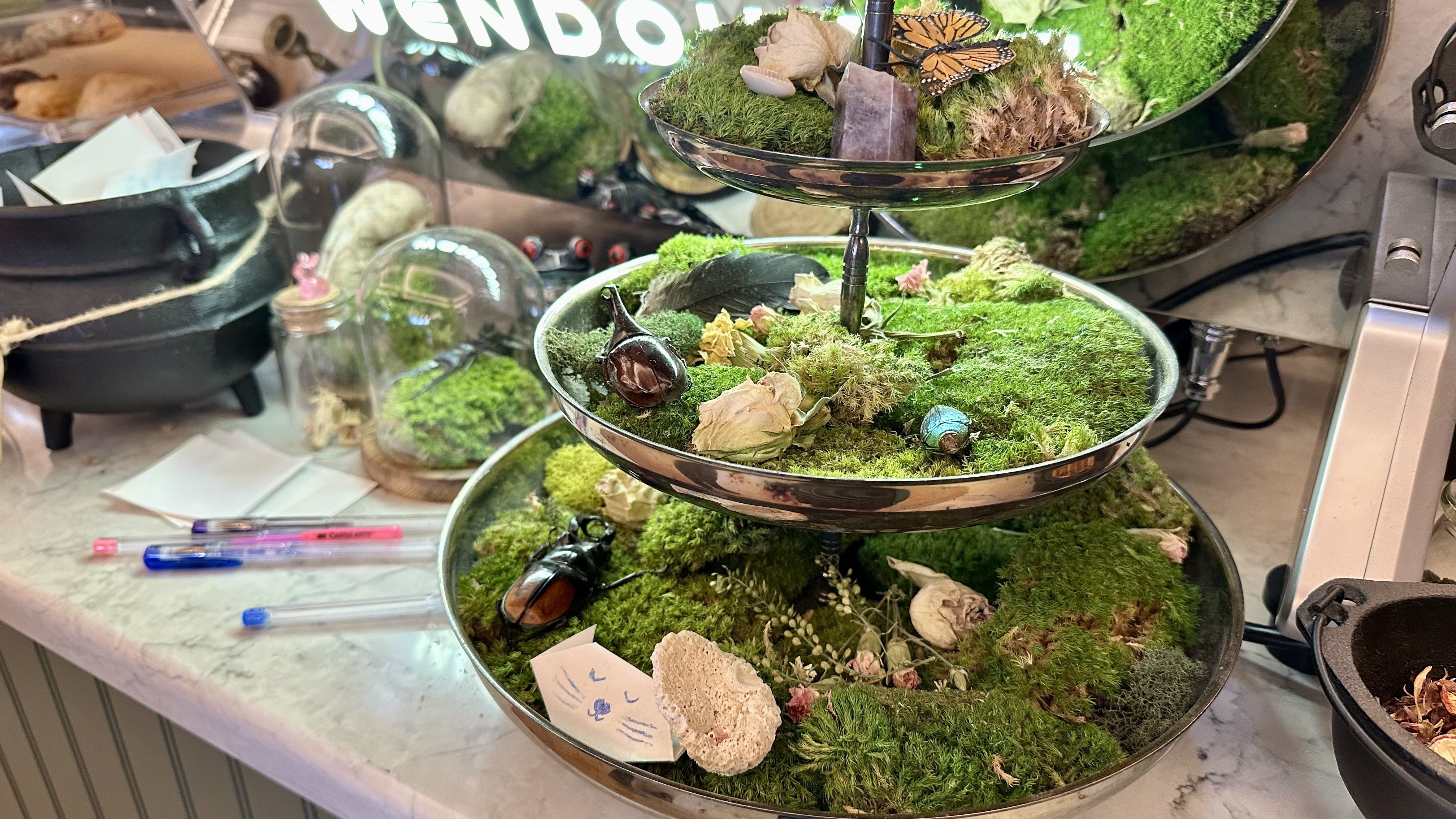 Three-tier metal moss display with small natural pieces and butterfly decorations, resting on a cluttered desk; glass cloches, dried leaves, shells, and a neon sign blurred in back.