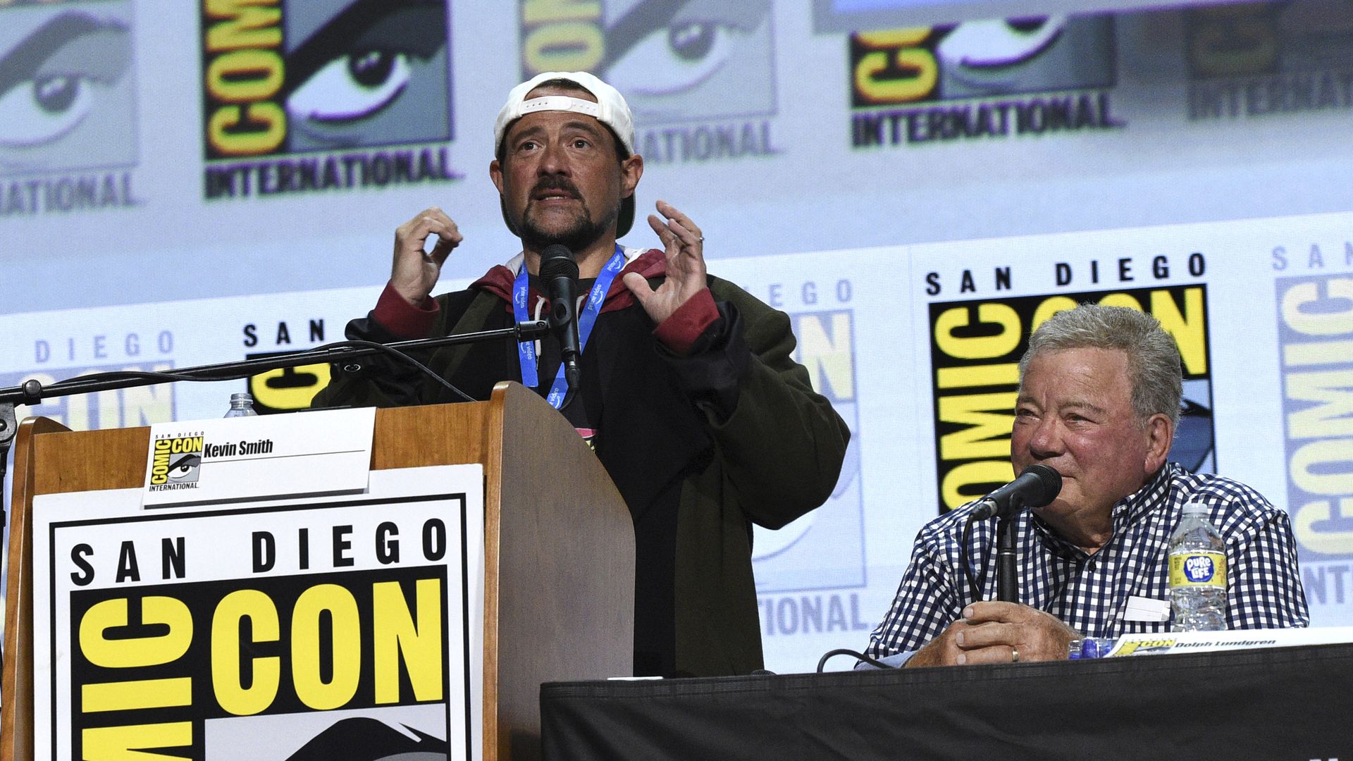 Kevin Smith speaks at a podium and William Shatner speaks into a microphone while seated