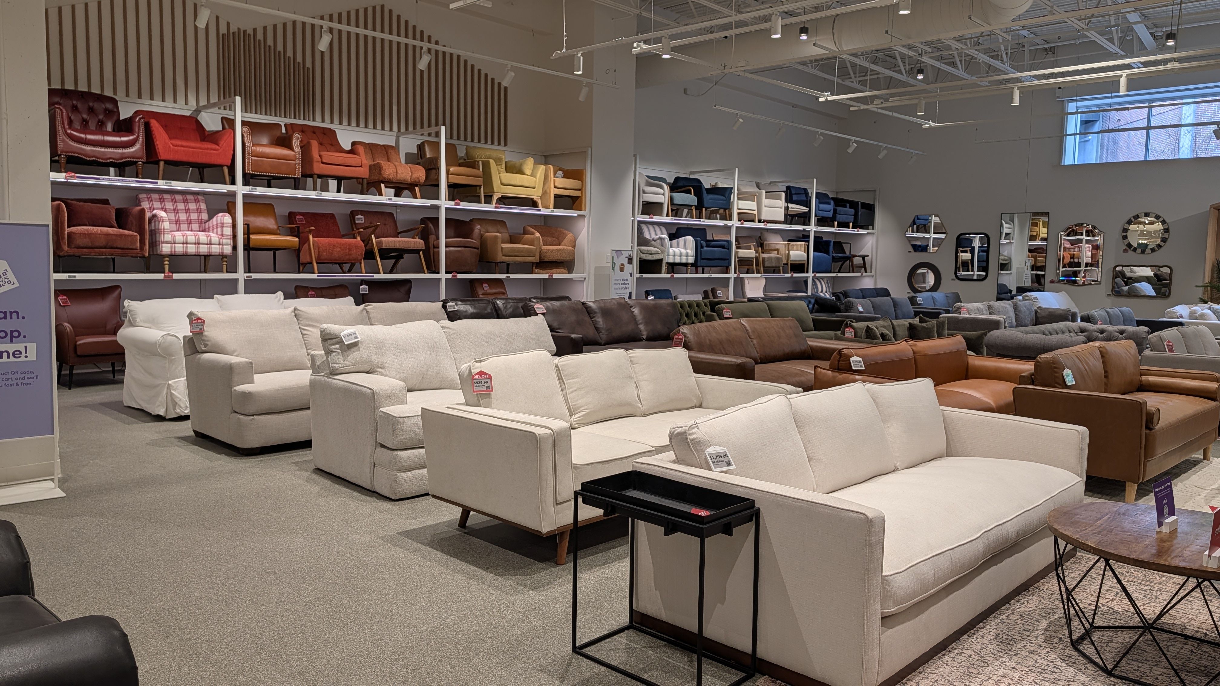 Interior of a furniture store showroom with rows of beige, white, and brown sofas. Wall shelves hold colorful chairs; bright track lighting and mirrors line the right wall.