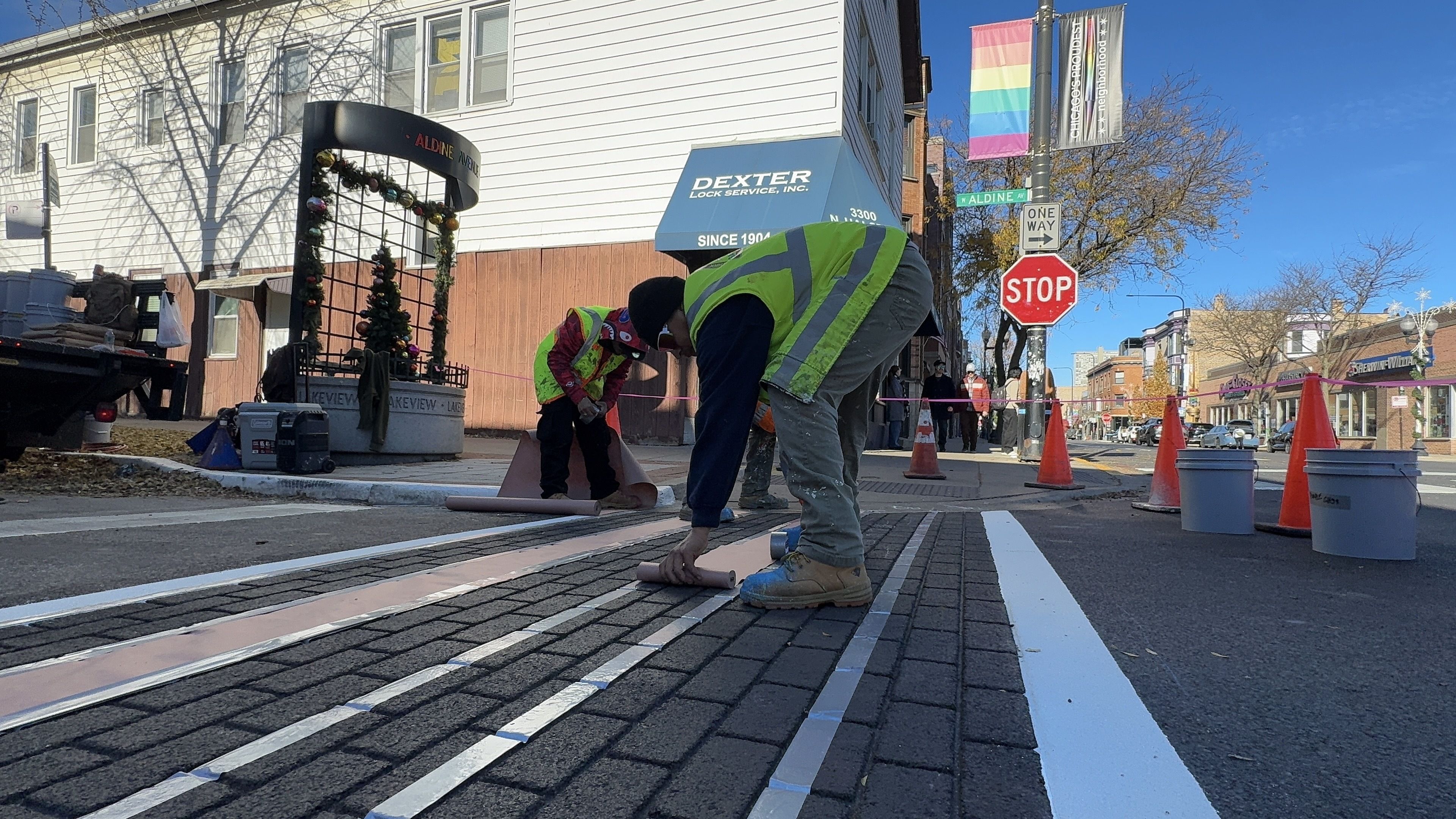 Two workers in neon safety vests install light pink strips on a brick-patterned street under a clear blue sky near a stop sign and a rainbow pride flag on Aldine Avenue.