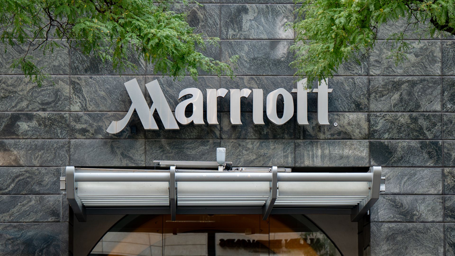 White Marriott sign on a dark gray stone wall, partially obscured by overhanging green tree branches; a glass entrance with a metal canopy visible at the bottom.