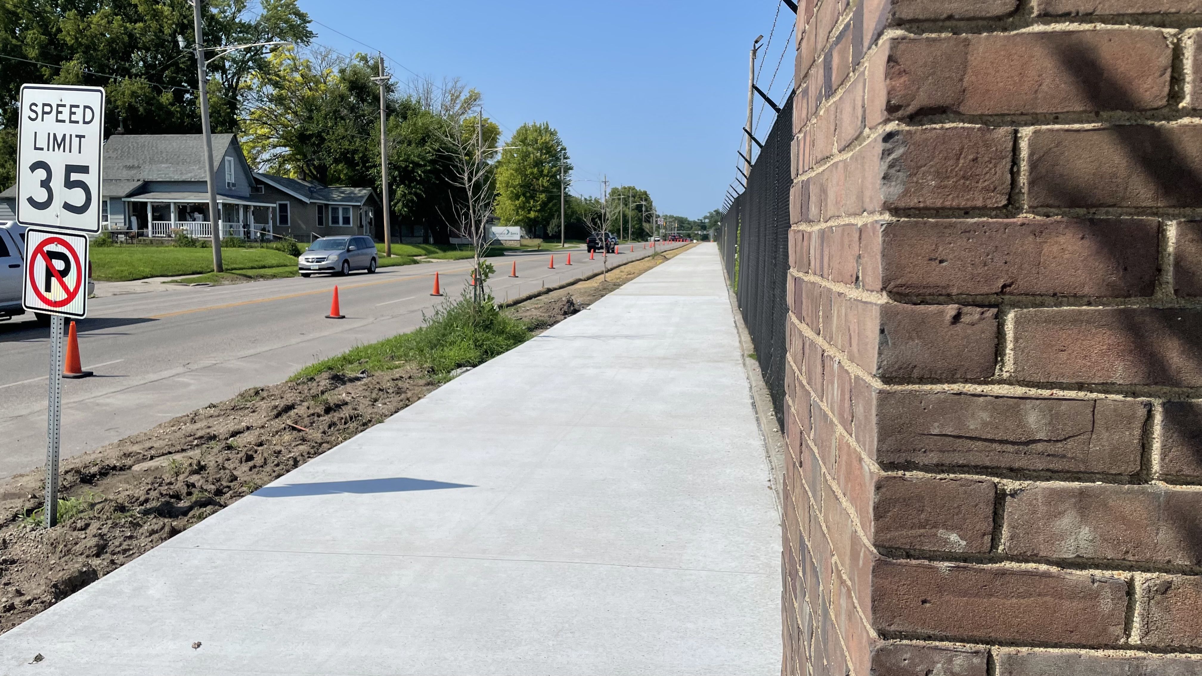 Long concrete sidewalk along a brick wall and black fence, next to a street with orange traffic cones, grassy area, a speed limit 35 sign, and residential houses under a clear blue sky.