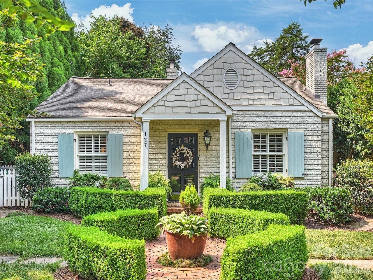 Hot homes. Charlotte houses for sale. A charming single-story brick cottage with light gray shingles on the gable, blue window shutters, a black front door with a wreath, and manicured boxwood hedges along a brick path to the entrance.