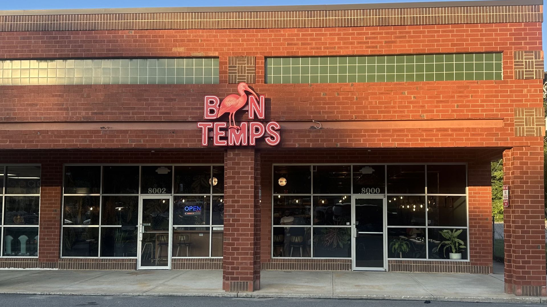 Red brick storefront with a neon pink sign reading BN TEMPS and a pink flamingo perched on the letters. Large green-tiled windows, decorative brick accents, and a blue sky above.