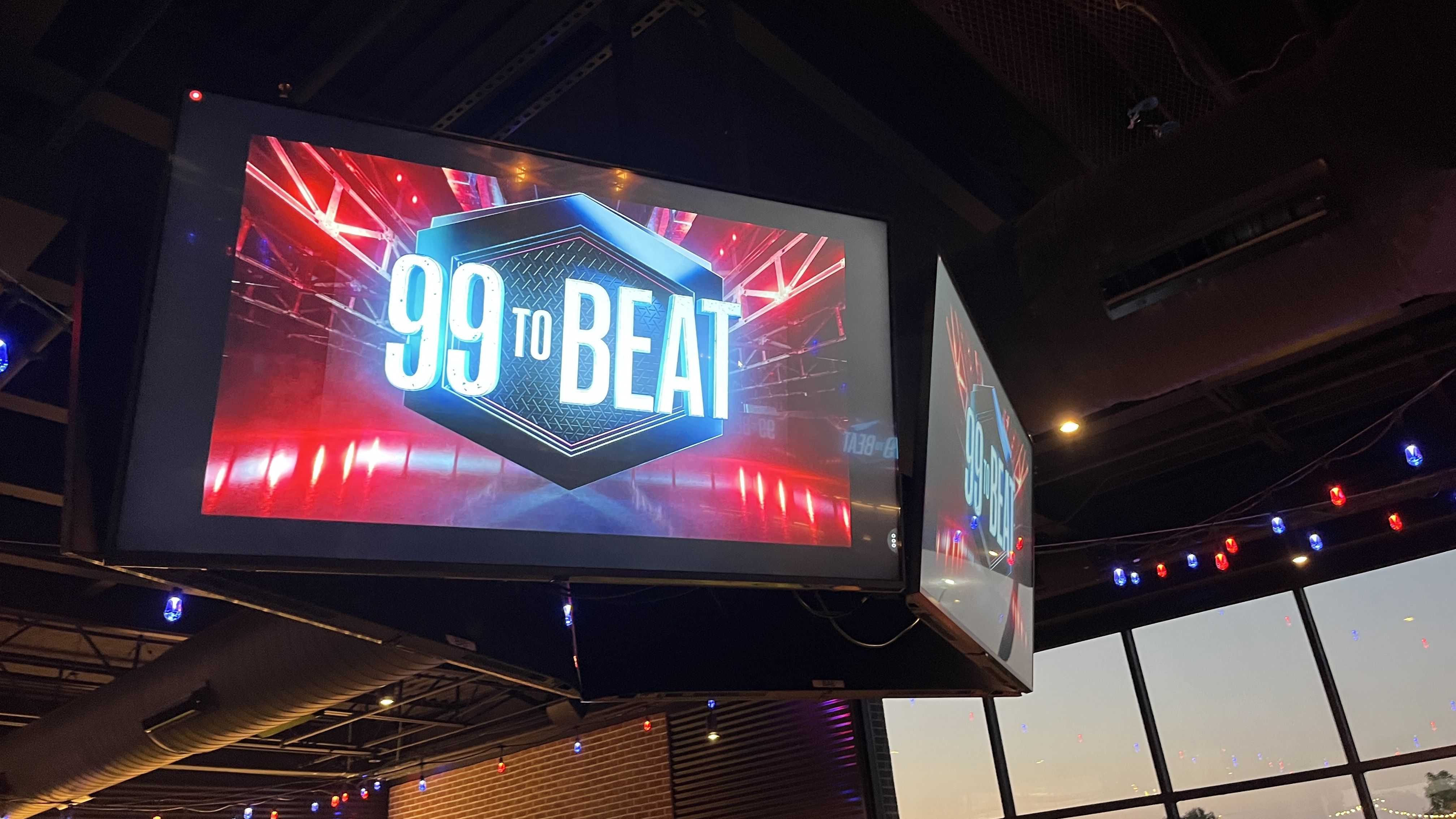 Indoor scene with a large screen displaying a hexagonal logo reading "99 TO BEAT" in white text on a blue and red illuminated background, surrounded by string lights in red and blue.