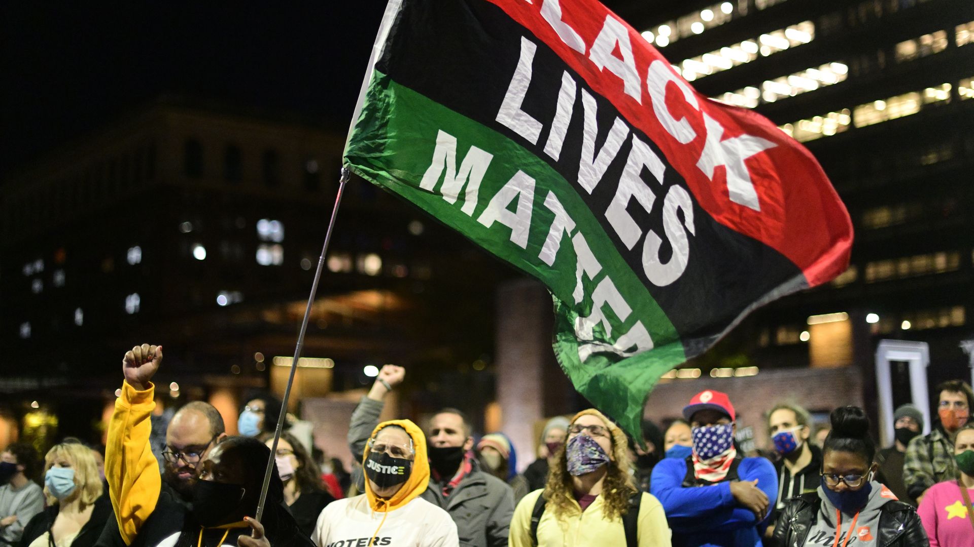 A Black Lives Matter flag hangs above a group of people walking in the street at night, with a city building behind them