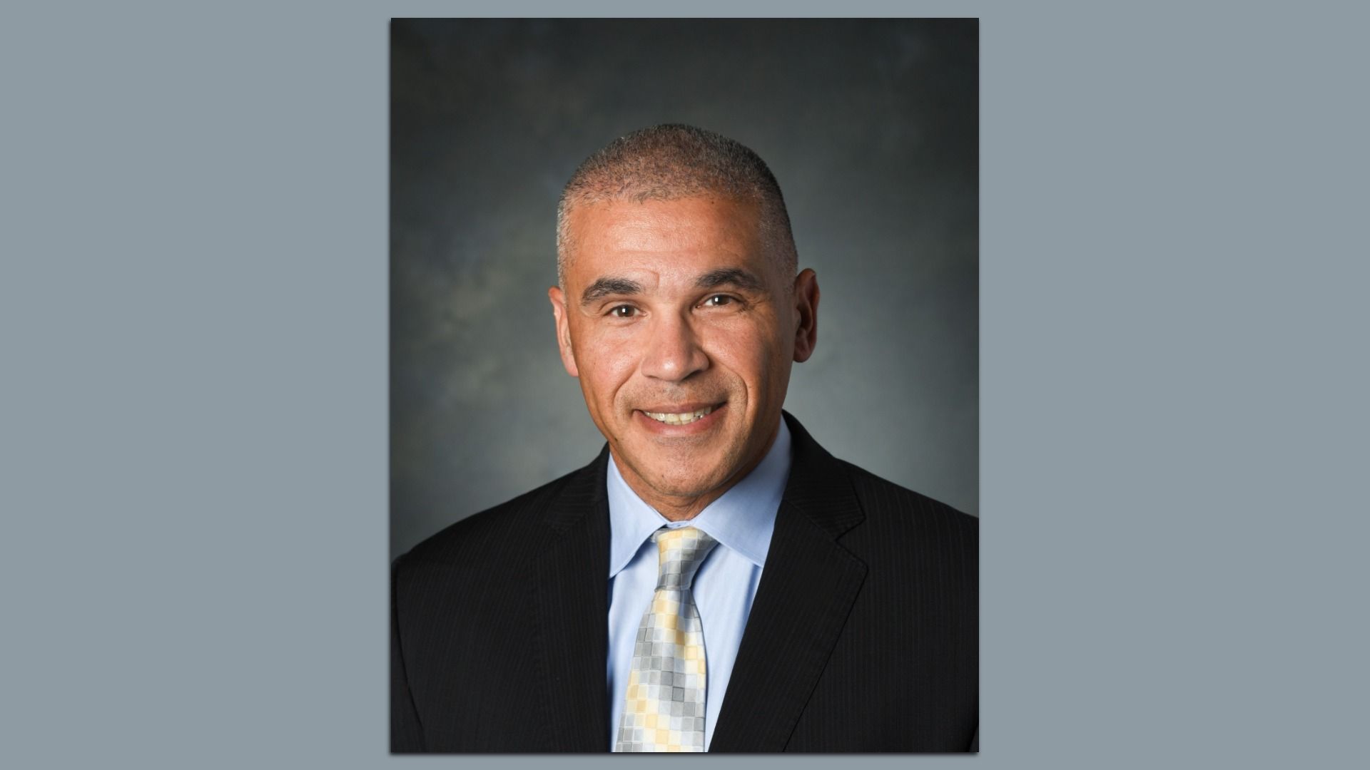 Portrait of a smiling middle-aged man with short gray hair, wearing a black suit, light blue shirt, and patterned gray and yellow tie, against a dark gray background.