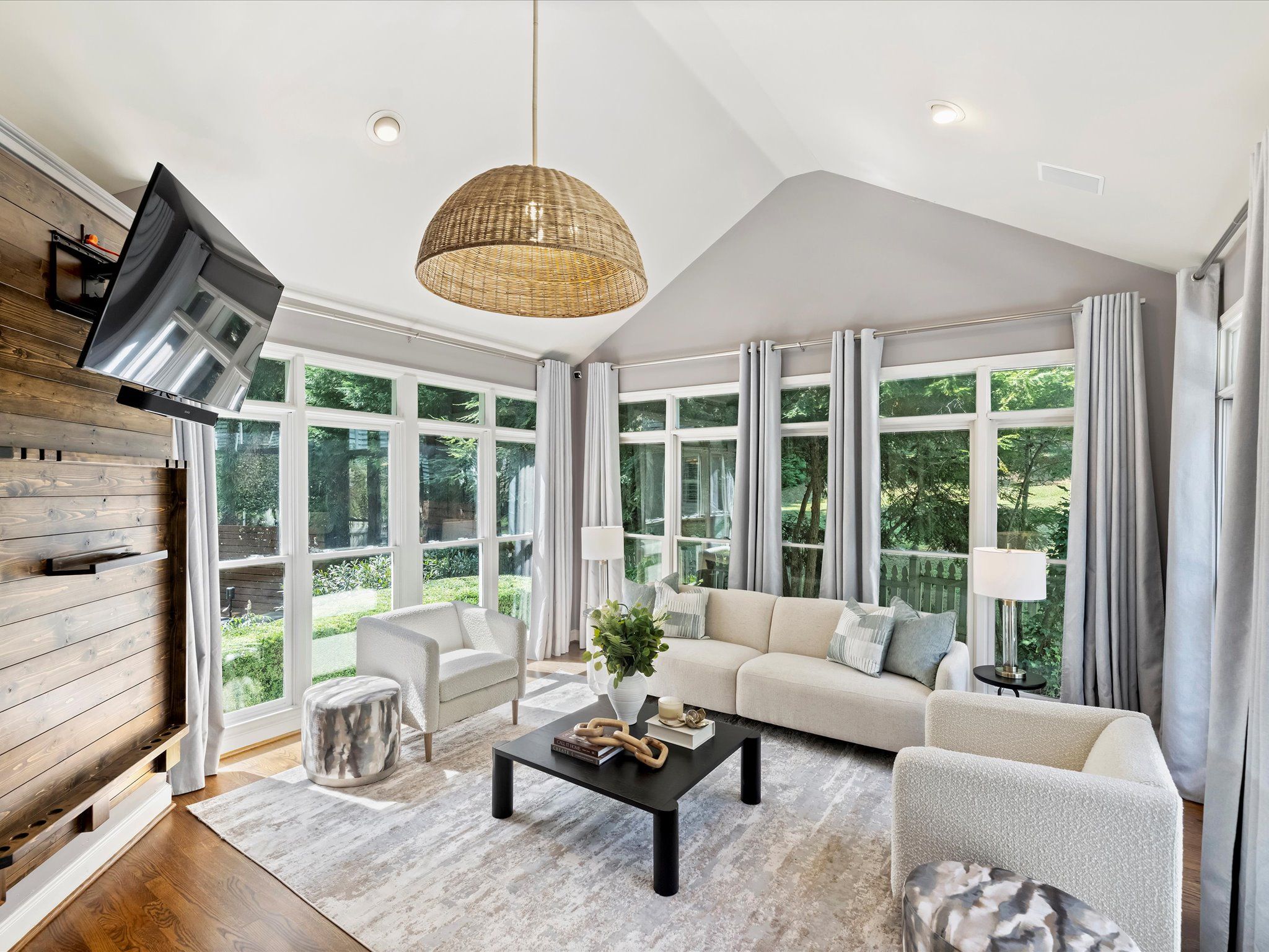 Bright living room with large windows, white curtains, beige sofa, two white armchairs, black coffee table with decor, wood accent wall with mounted TV, and woven pendant light.