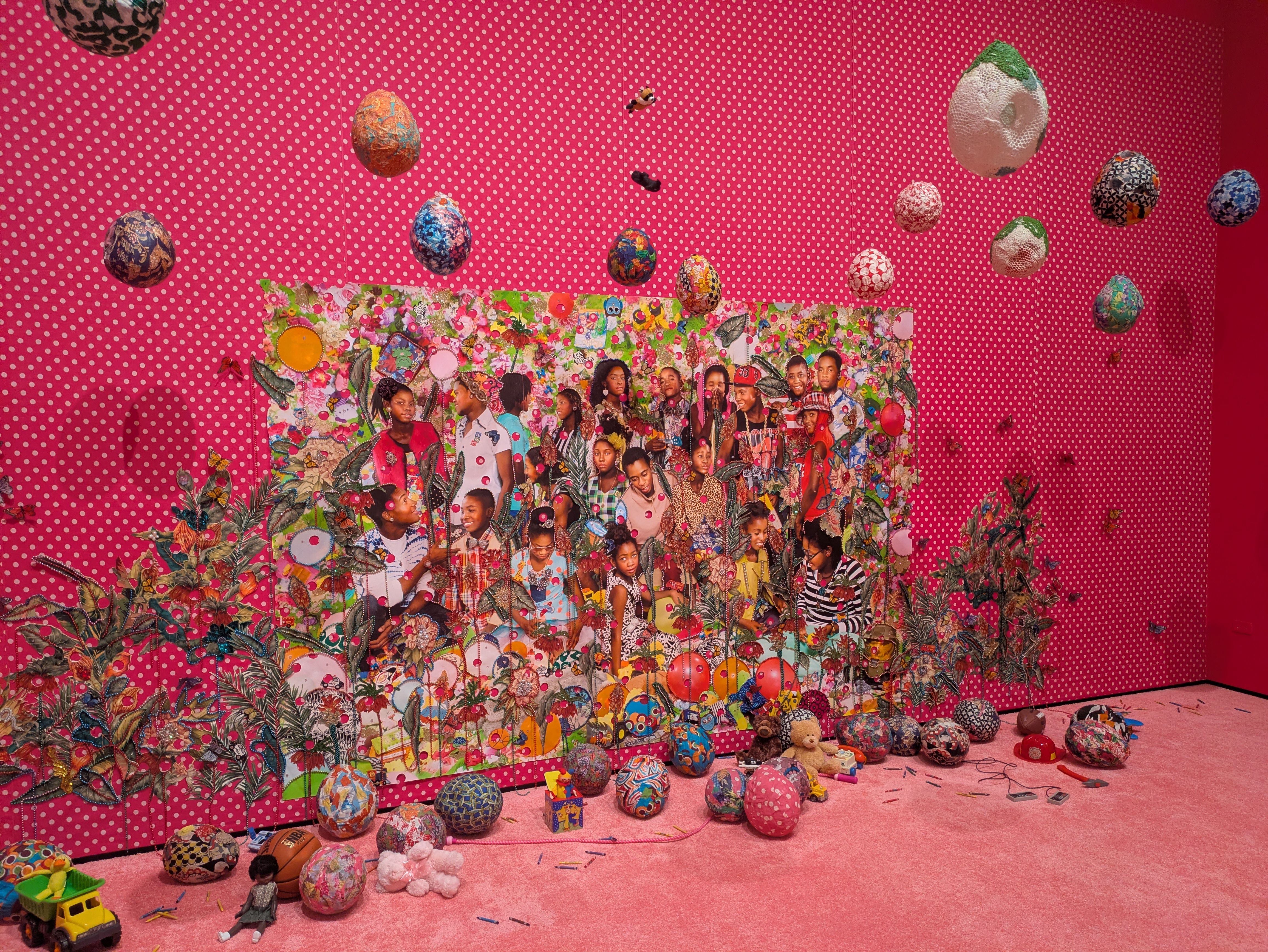 Pink wall with white polka dots, colorful decorated eggs hanging and scattered on the floor; a collage of diverse people at the bottom and several toys on a pink carpet.