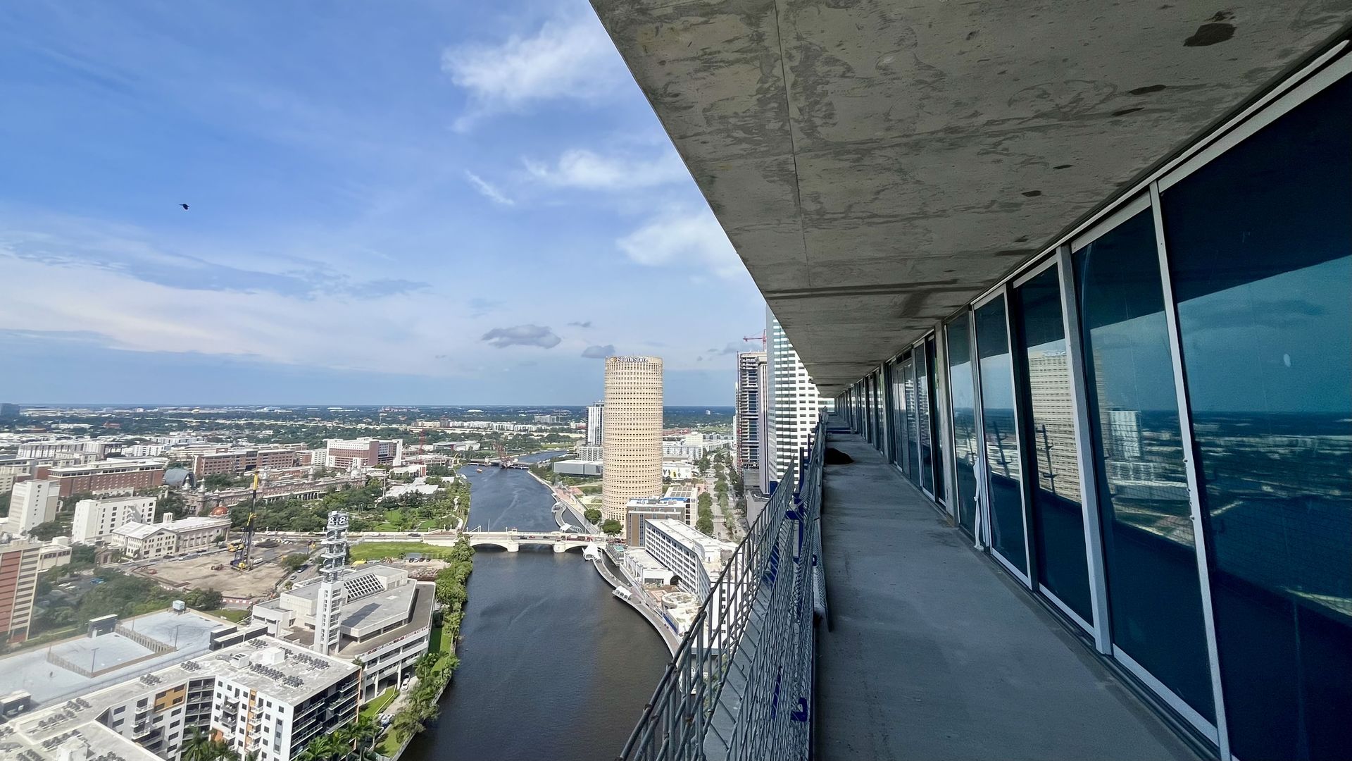 View from a high balcony along a modern skyscraper: glass windows on the right, concrete ceiling above, a city with a river and bridge below, and a lone bird in the blue sky.