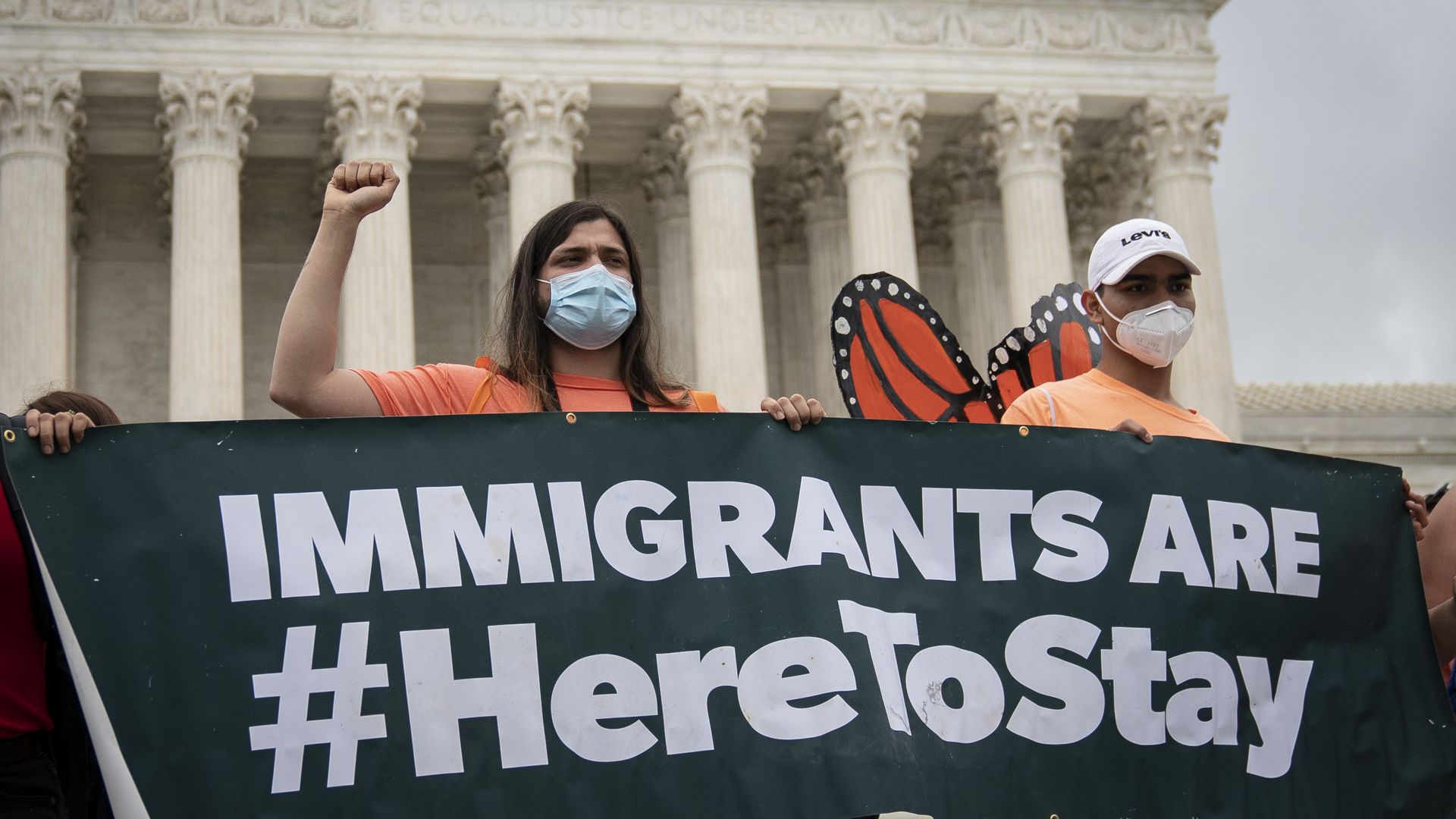 Photo of two masked people standing in front of the Supreme Court and holding a sign that says "Immigrants are #HereToStay"