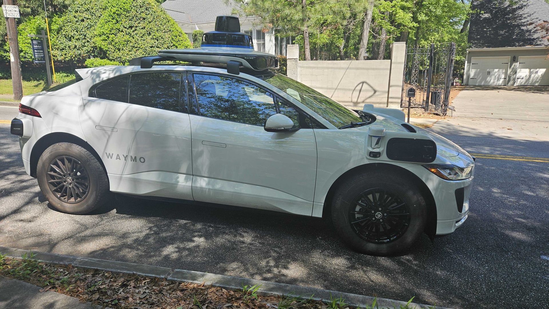 A photo of a white autonomous electric vehicle with spatial awareness equipment on the roof. The word "Waymo" is emblazoned on the rear of the vehicle. 