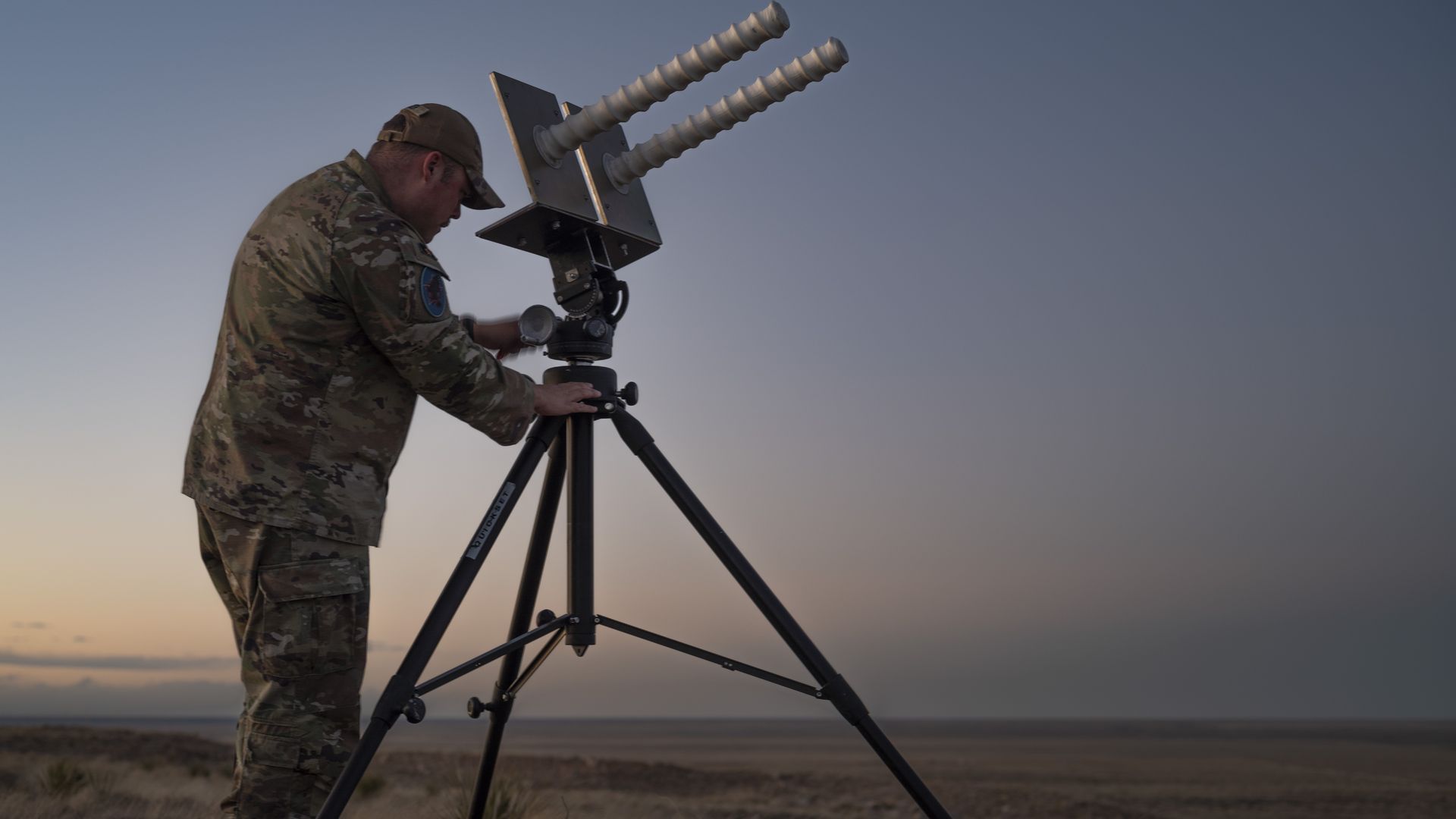 A soldier in camouflage adjusts a large tripod-mounted antenna with two ribbed insulators at sunset over a desert landscape.