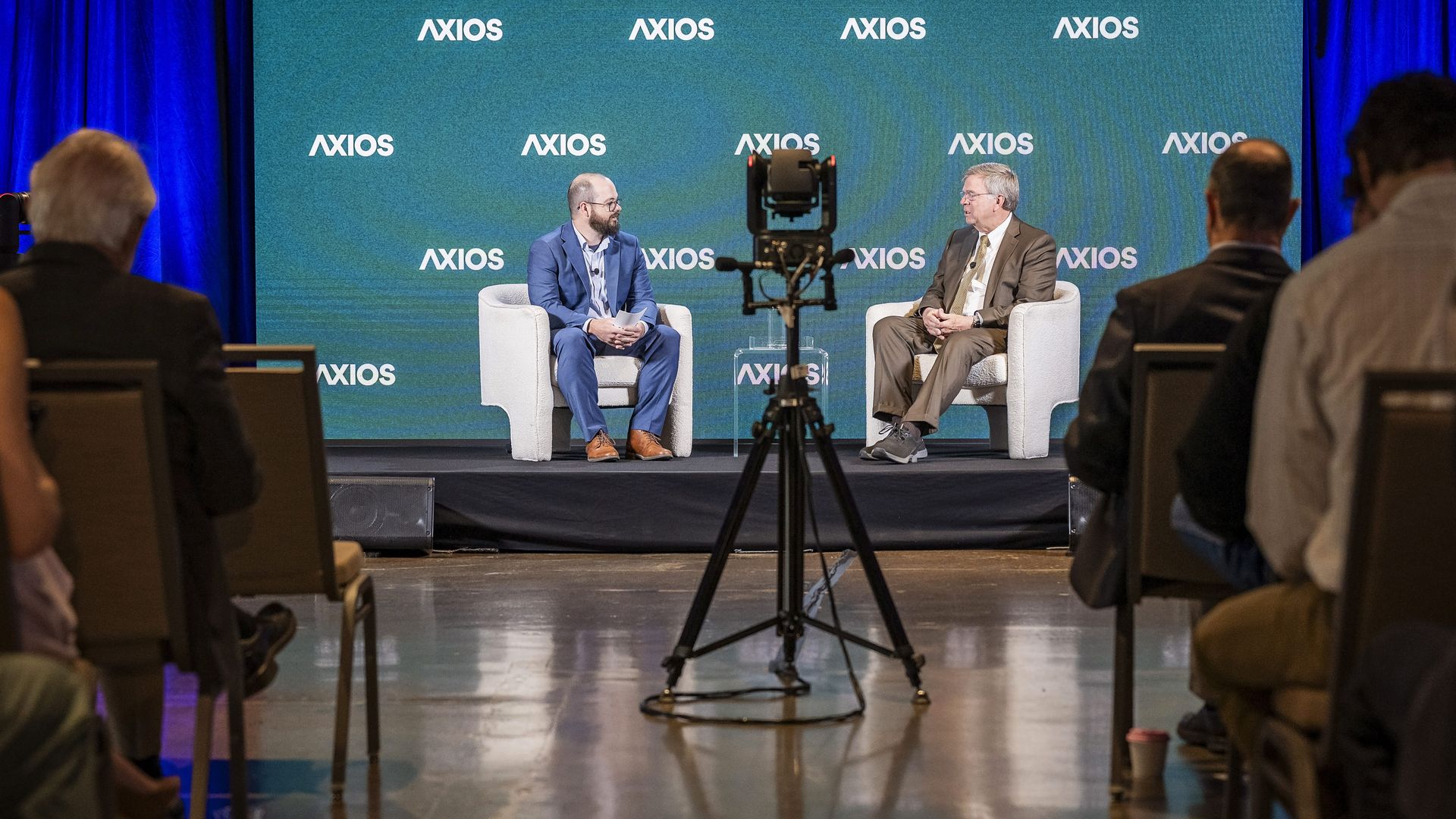 Two men in suits sit on a stage with a teal Axios backdrop, engaged in a talk-show discussion. A camera on a tripod is centered between them, while an audience sits in the foreground.