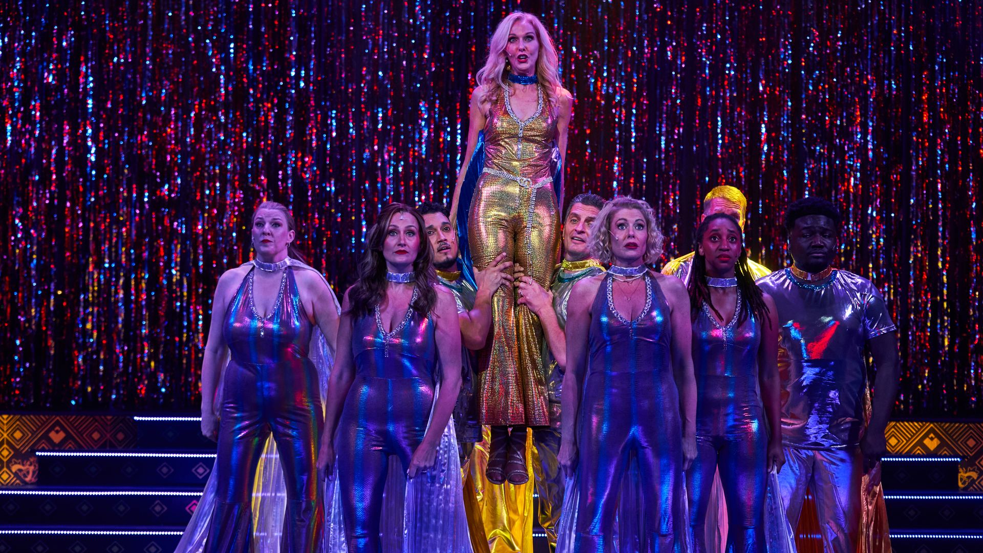 A lead performer in a gold metallic jumpsuit is lifted by dancers in silver outfits on a glittery, red-and-blue curtain stage.