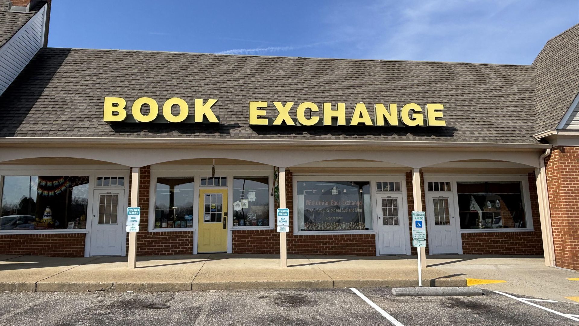 Front of a bookstore with a brown shingled roof and a large yellow sign reading "BOOK EXCHANGE". Brick storefront, white doors, big display windows, a yellow center door, and a clear blue sky.