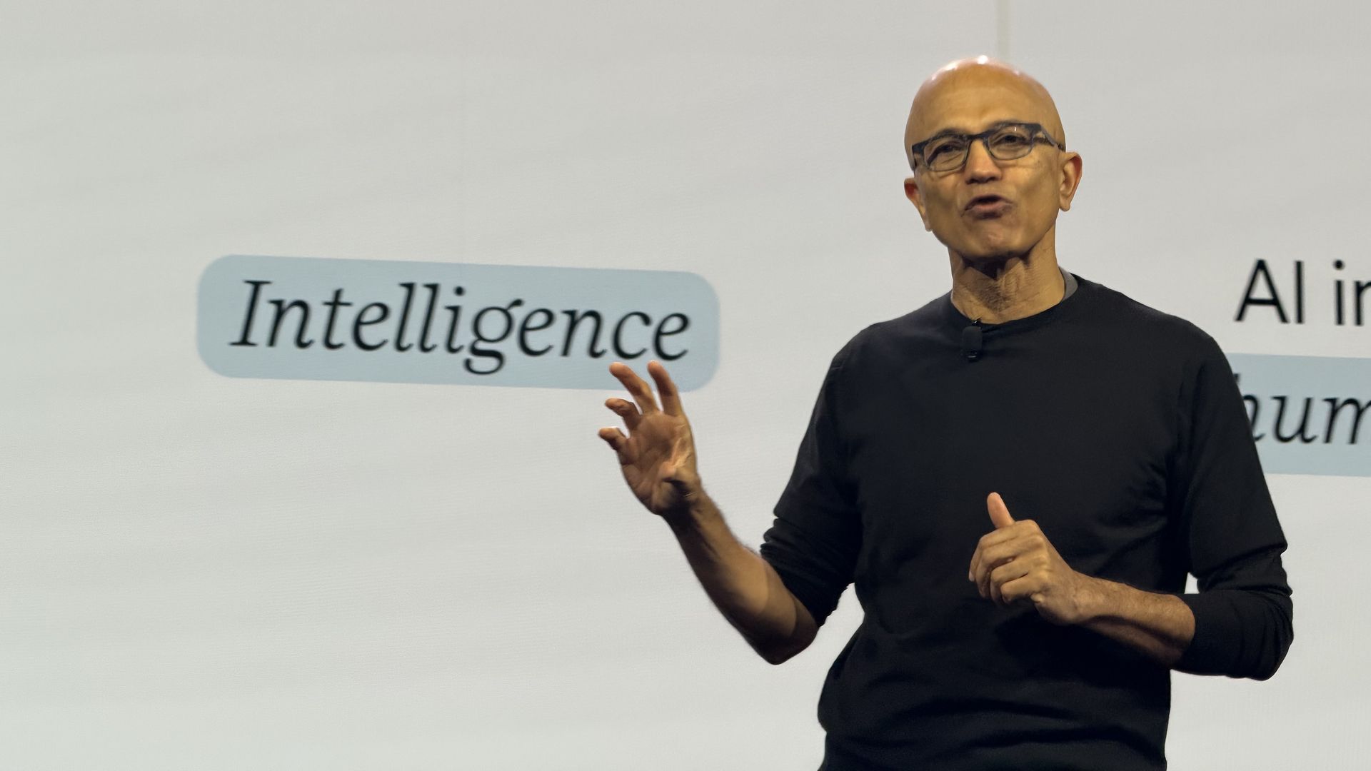 A speaker in a black shirt and glasses stands on stage, gesturing with his hands, as the word "Intelligence" appears on a screen in the background.