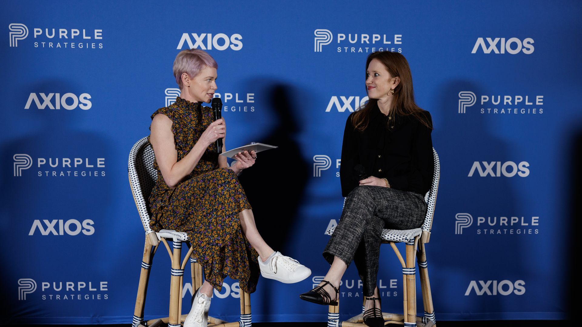 Two women seated on wooden stools in front of a blue backdrop with Purple Strategies and Axios logos, one in a floral dress holding a microphone and tablet, the other in black top and gray pants holding a microphone.