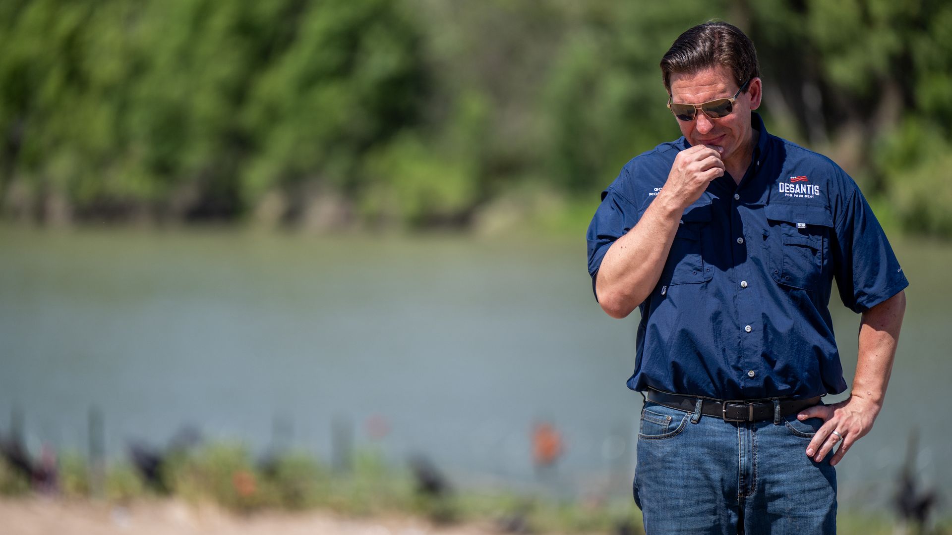 Ron DeSantis, a man in a blue shirt and blue jeans, stands near a river. He is positioned on the right side of the picture. He is rubbing his chin and wearing sunglasses.