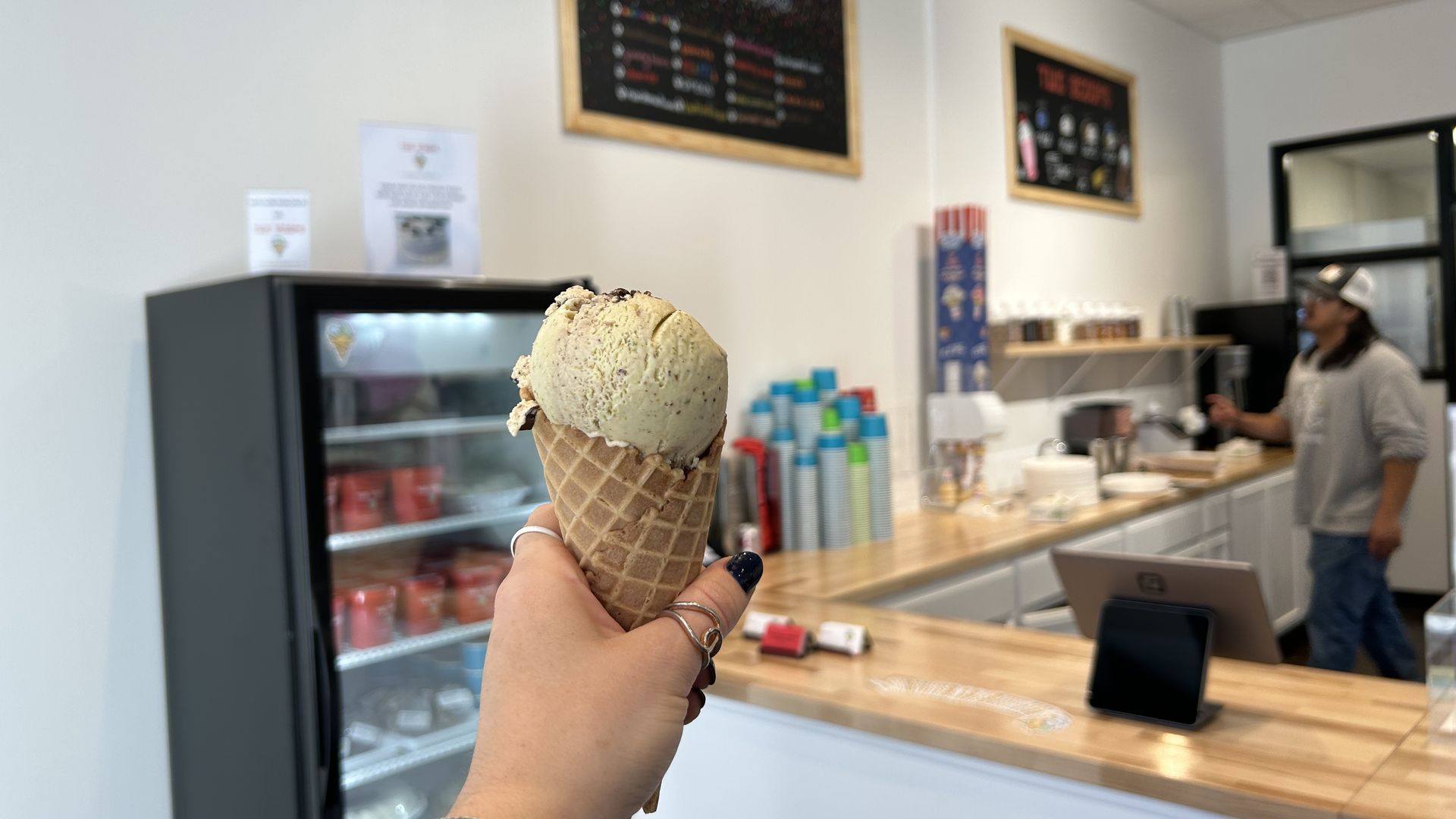 Hand holding a waffle cone with light-colored ice cream inside an ice cream shop, showing counter, a man in a cap, cones, and menu boards in the background.