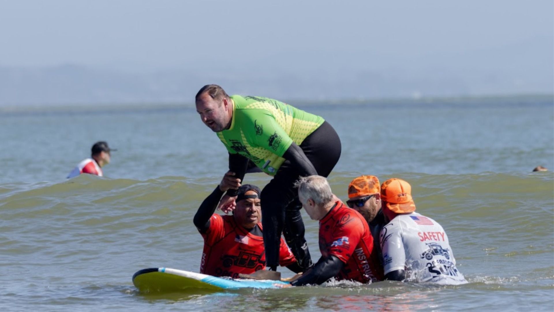 A man in a green wetsuit is standing on a surfboard, assisted by four men in red and white wetsuits with orange hats, in calm ocean water.