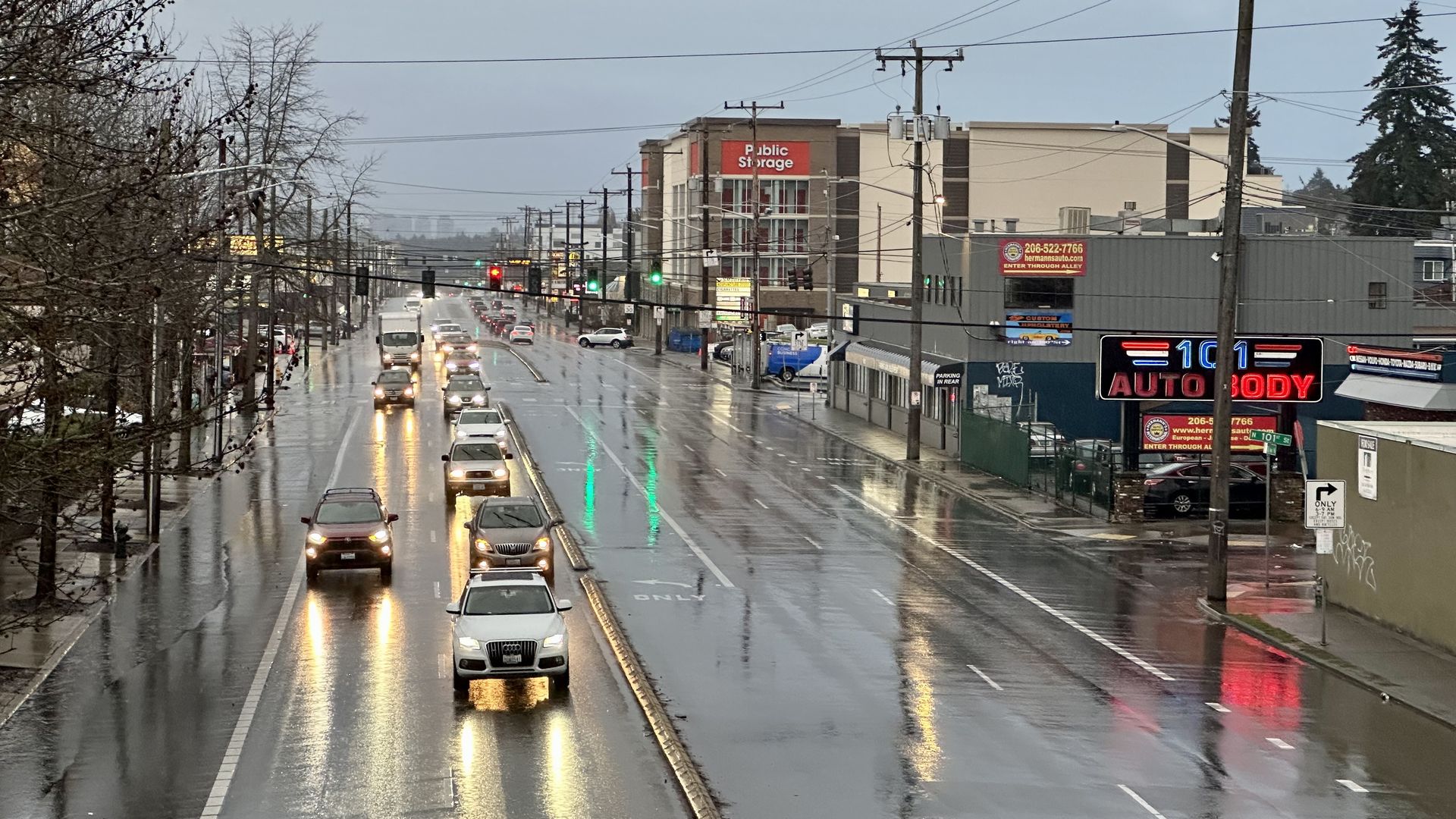 A wet  six-lane roadway with cars driving toward the camera and an auto store and a public storage facility visible on the right side of the highway.