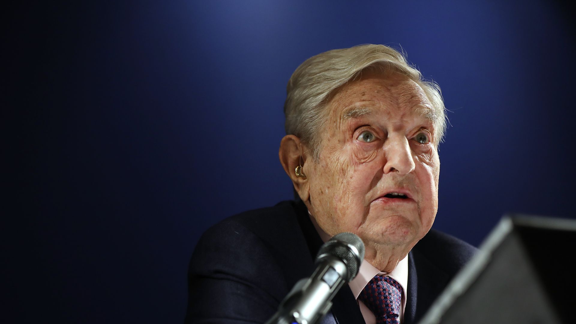 Billionaire and philanthropist George Soros speaks at the World Economic Forum, wearing a navy suit, along with a red and blue tie. 