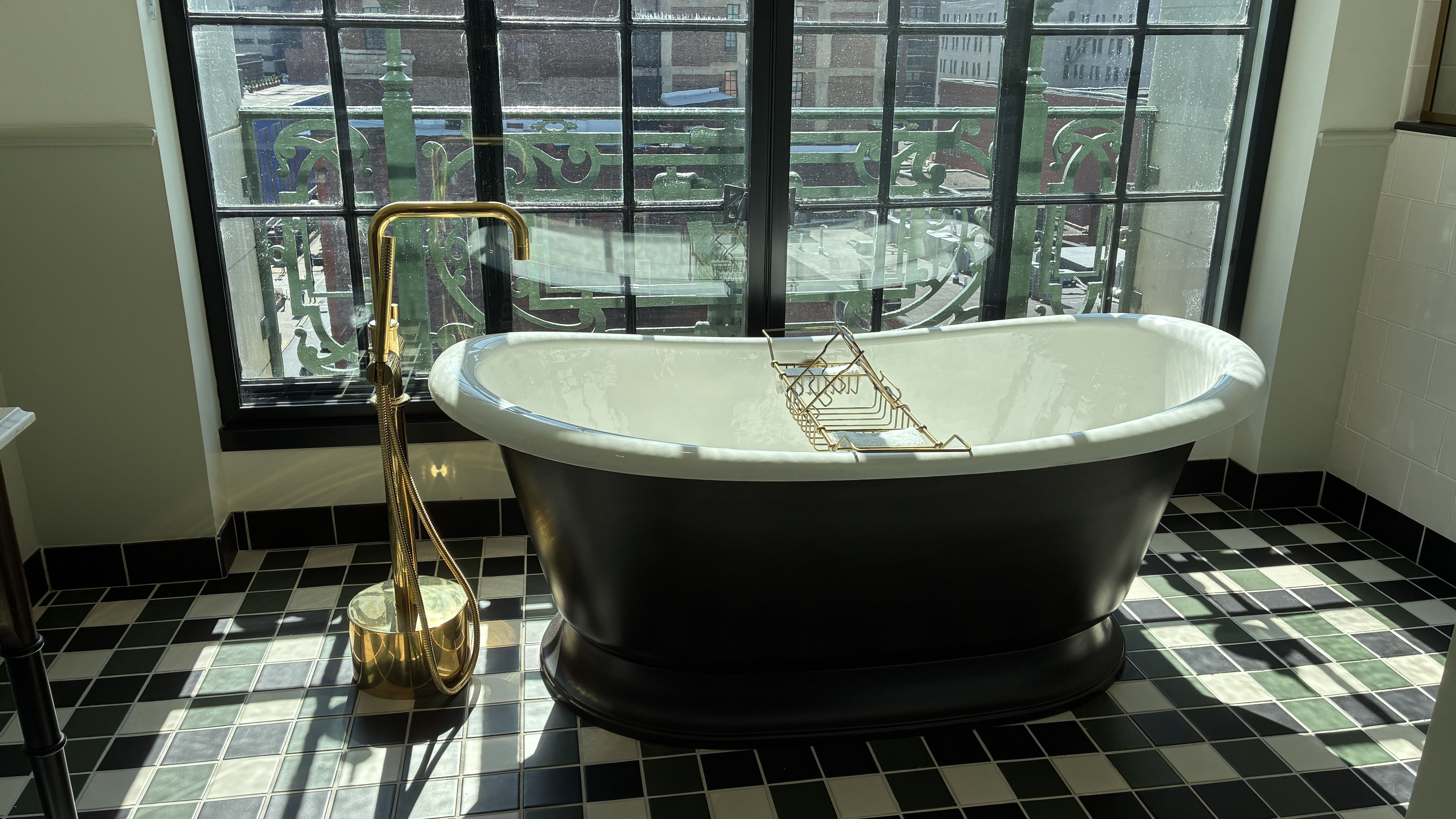 Interior view of a vintage freestanding black tub with white interior, gold faucet and stand, on a black-and-white checkered tile floor. A green wrought-iron balcony window frames the outside.