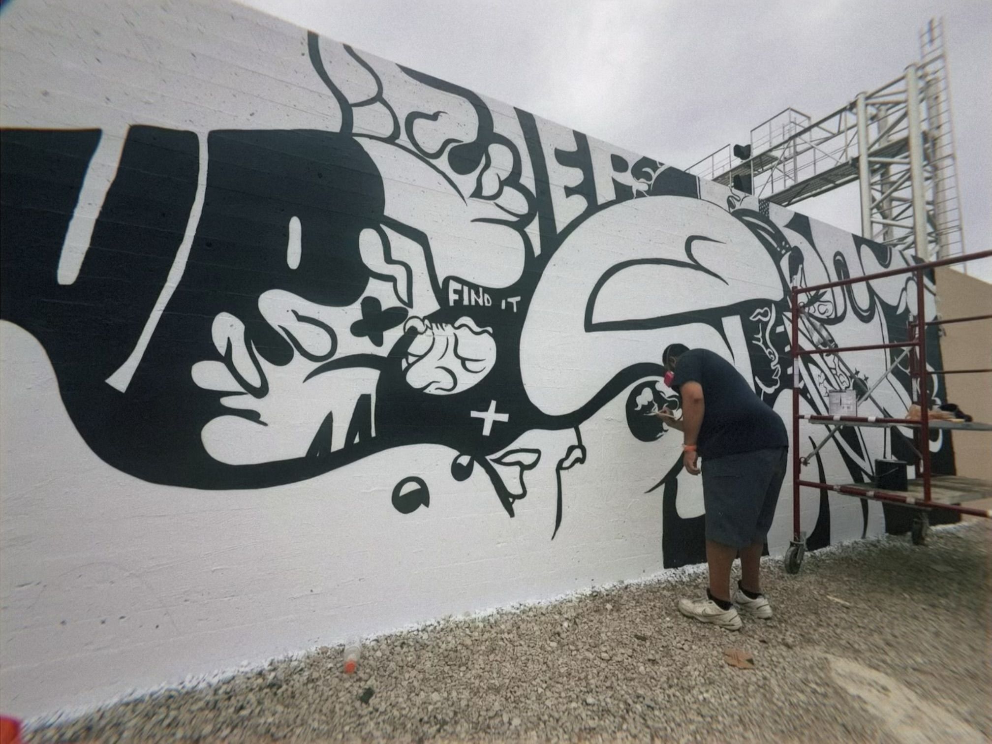 Person painting a large black and white abstract mural on a white brick wall outdoors, featuring organic shapes and the words "FIND IT". Scaffolding and gravel ground visible.