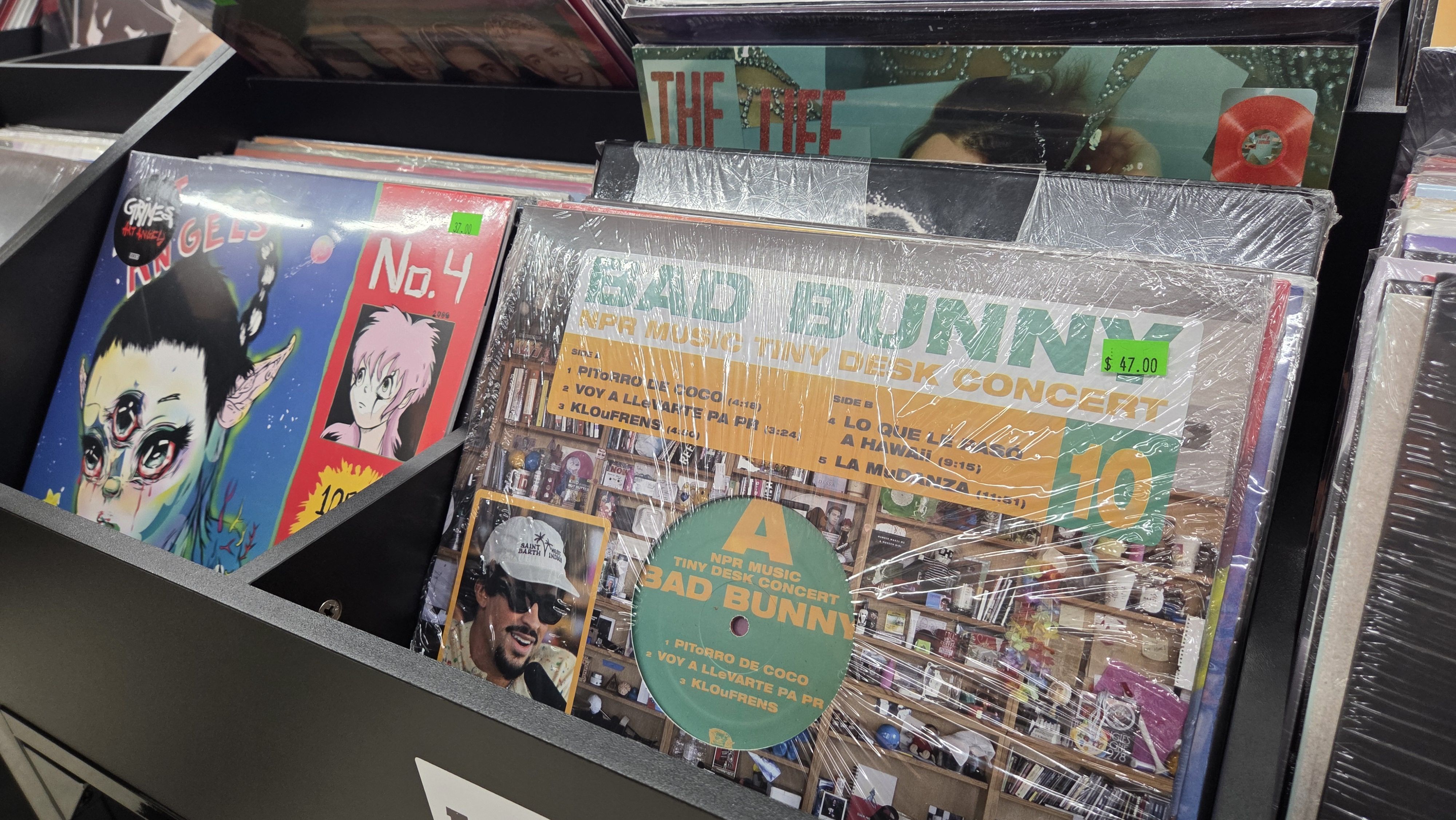 Vinyl records in a store bin, featuring "BAD BUNNY NPR Music Tiny Desk Concert" with price $47 and colorful album covers including one titled "ANGELS No. 4" with illustrated faces.