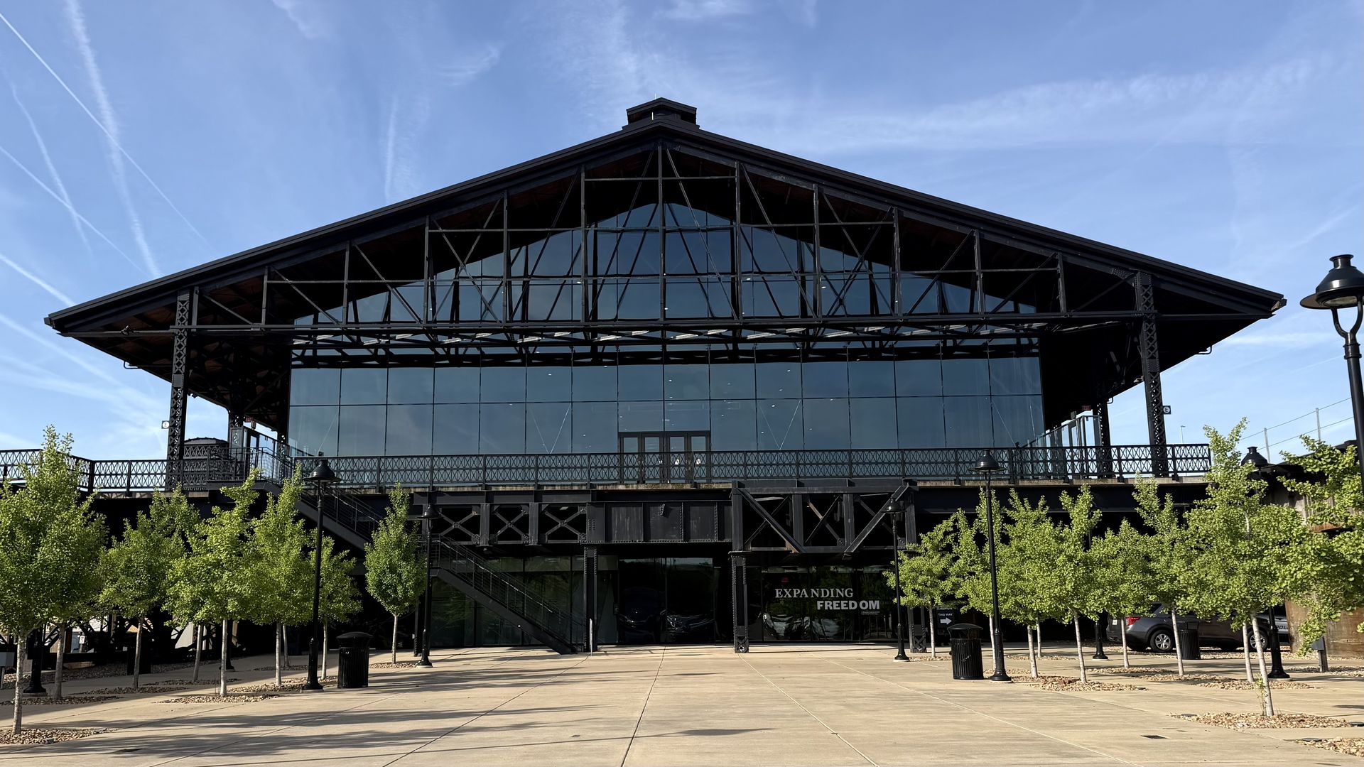 A large, modern black metal-framed building with a triangular roof and glass façade. Exterior shows iron trusses, a raised balcony, and a wide plaza with small trees and lampposts under a blue sky.