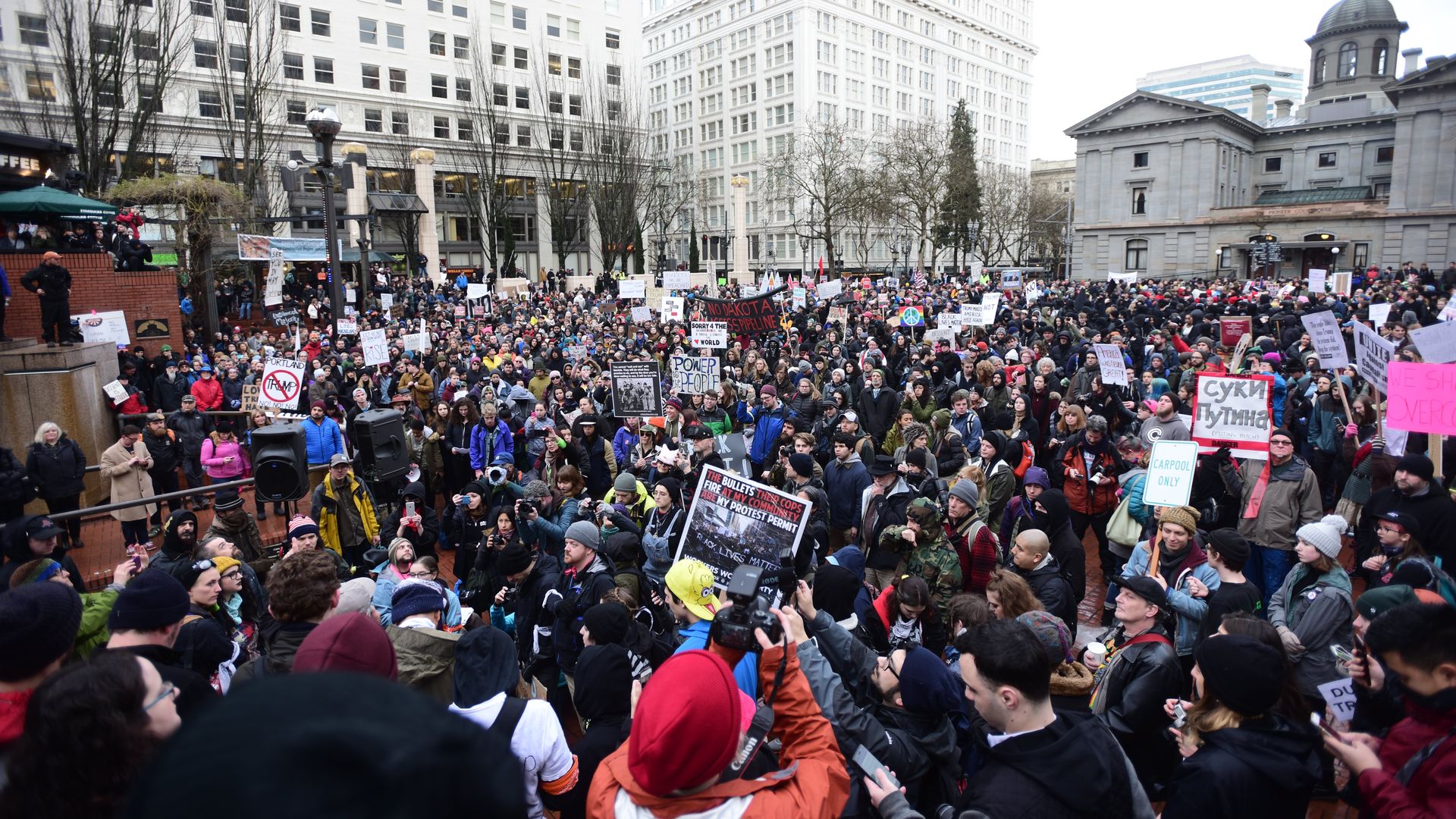 Large crowd of protesters in jackets and hats holding various signs in a city square with tall buildings and bare trees in the background on a cloudy day.