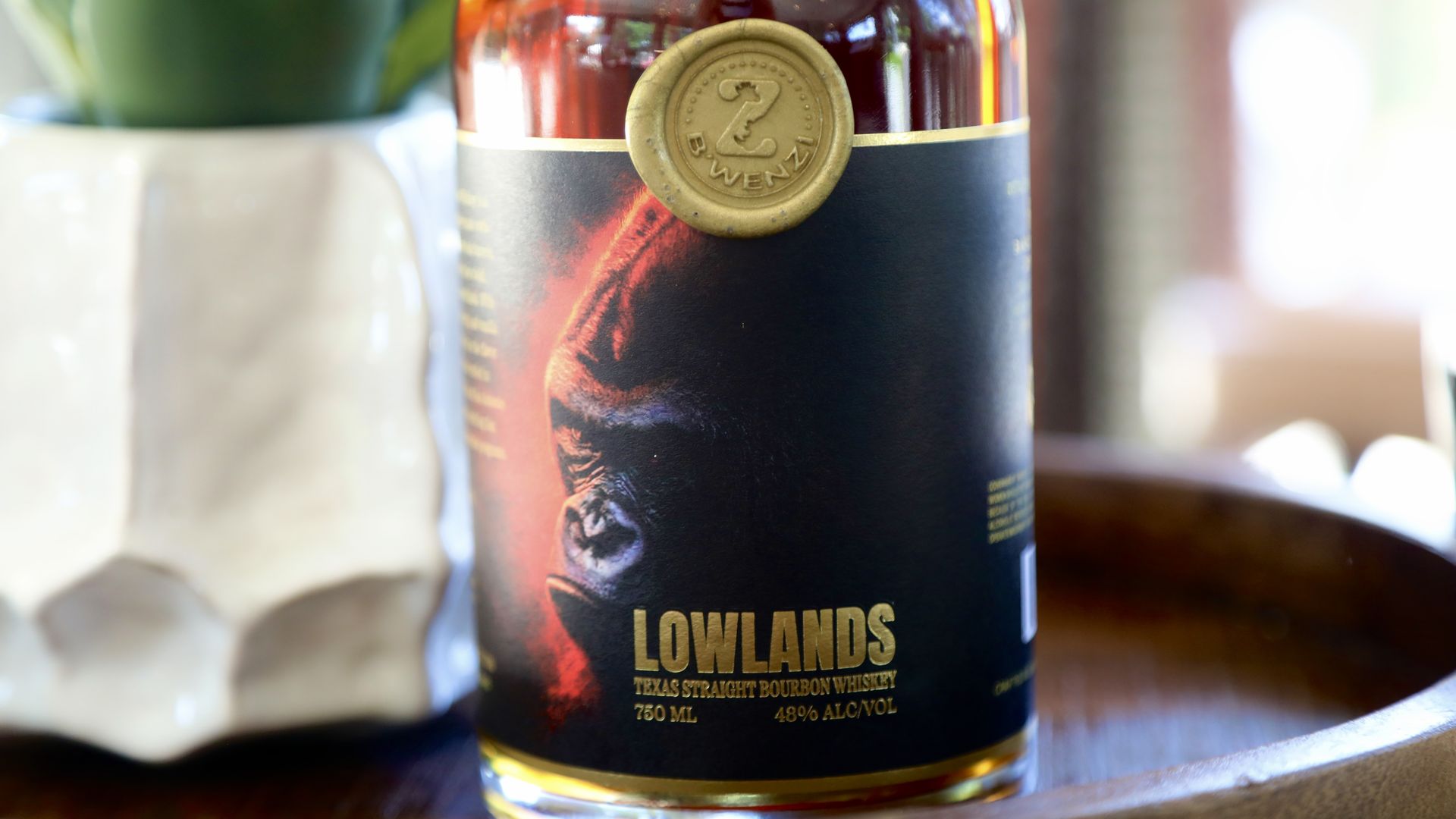 A close-up of a bourbon whiskey bottle that reads "LOWLANDS" at the bottom, sitting on a wooden tray with a blurred green plant in the background. Ranger Creek's B'Wenzi Lowlands bourbon is made exclusively for the San Antonio Zoo. 