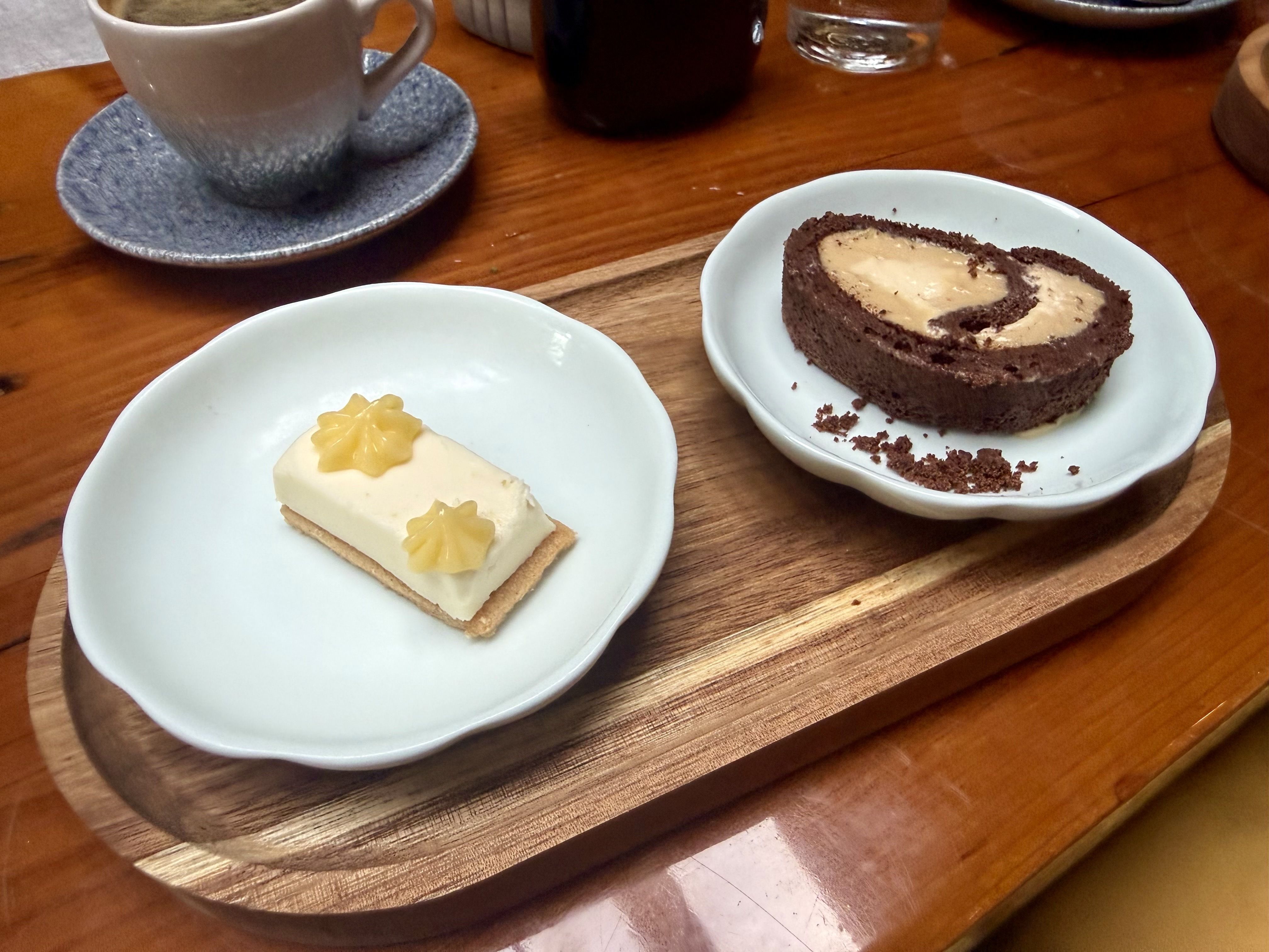 Chocolate roll cake with creamy filling on a white plate, crumbs around; a small cream-topped dessert with yellow flowers on a second plate; coffee cup and glass on a warm wooden table.