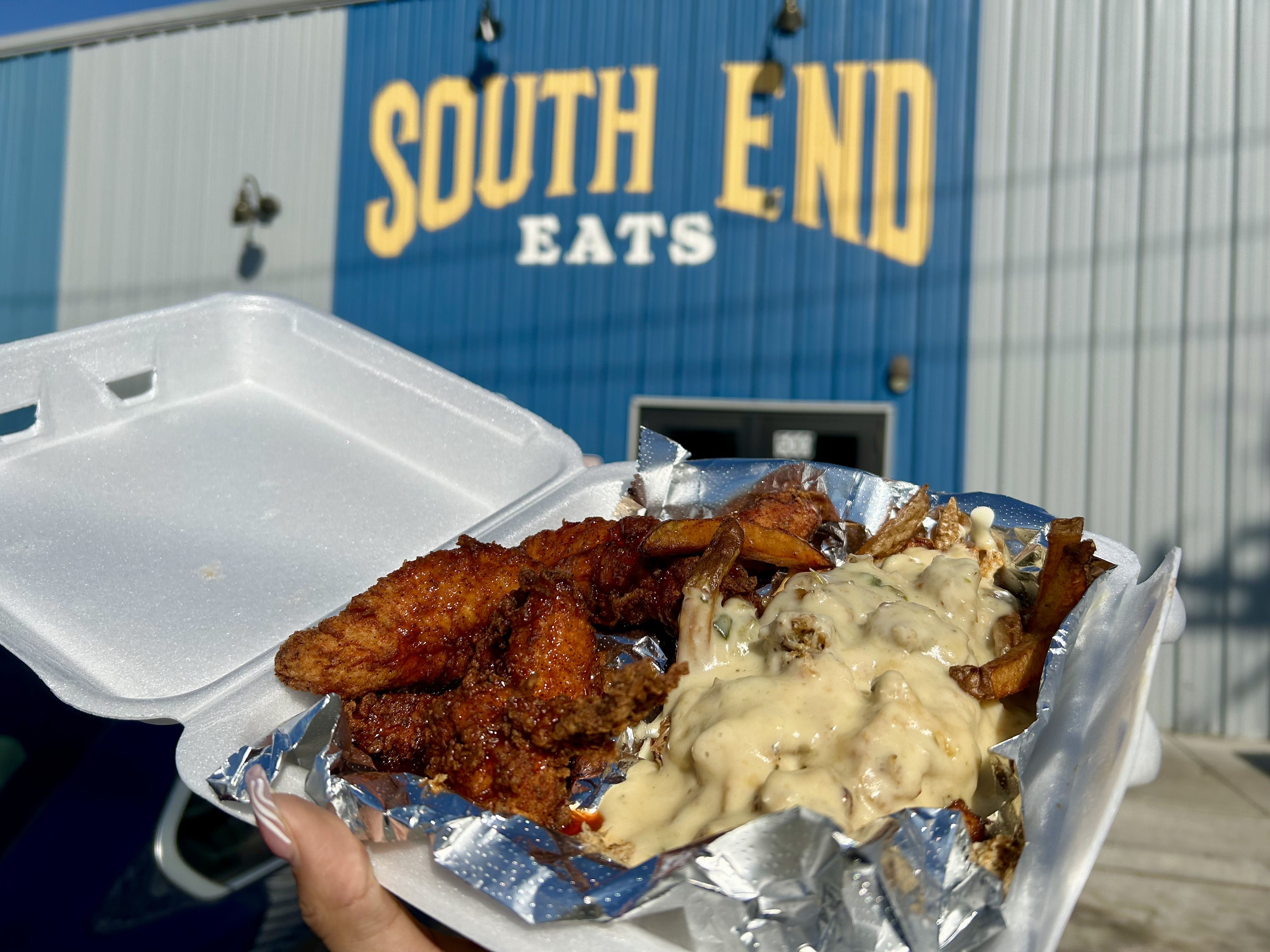 Takeout container with spicy fried chicken and fries covered in creamy gravy, held outside a blue building with large yellow letters reading "SOUTH END EATS".