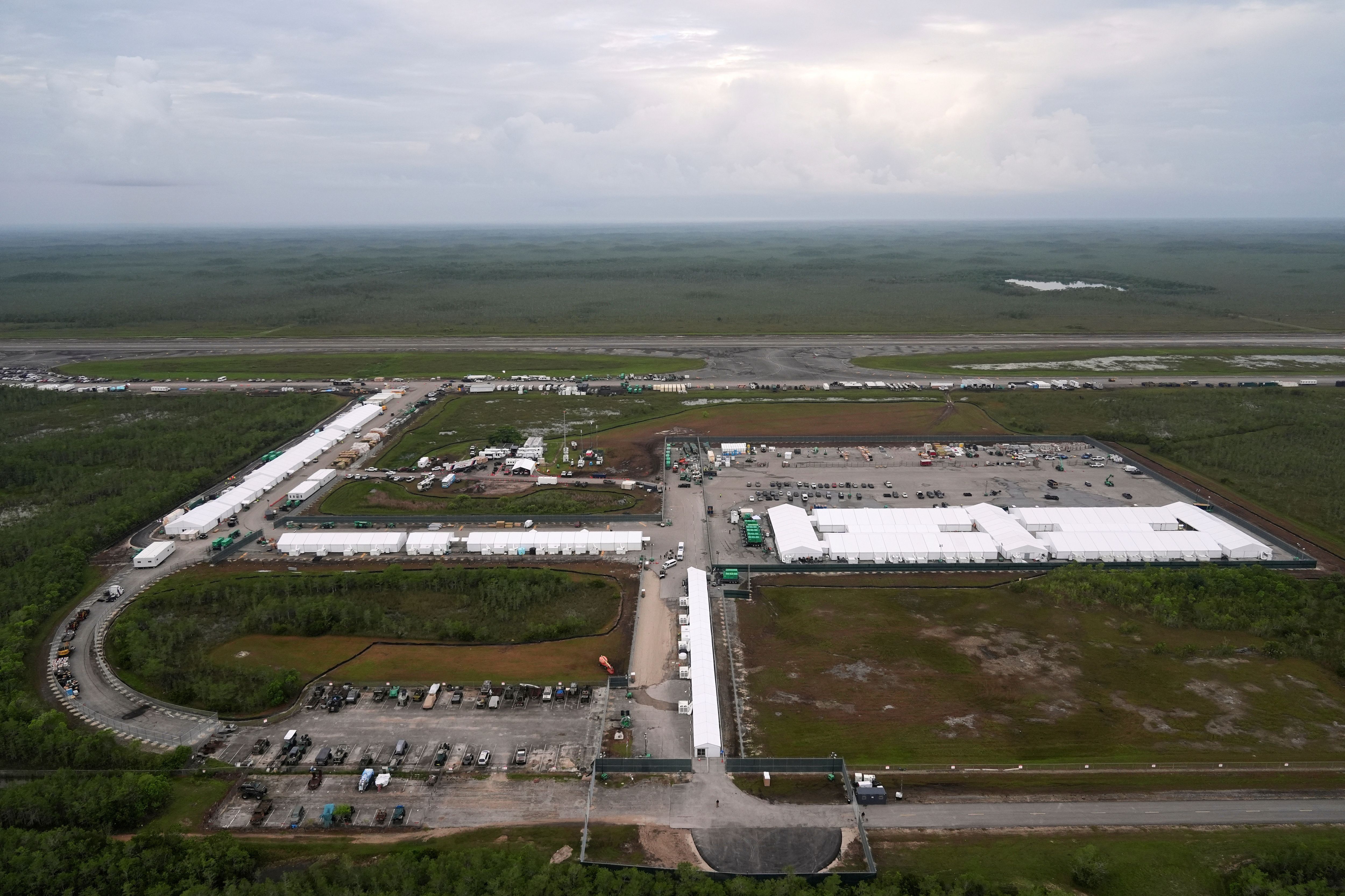 Aerial view of a large facility with numerous white tents and parked vehicles, surrounded by green landscape under a cloudy sky.